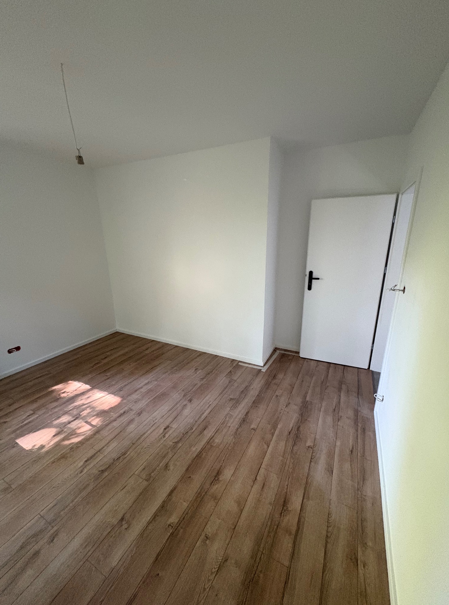 Jasne wnętrze z drewnianą podłogą, pomalowanymi na biało ścianami i drzwiami. Widoczne gniazdko elektryczne i kabel zwisający z sufitu. Wpadające światło słoneczne.