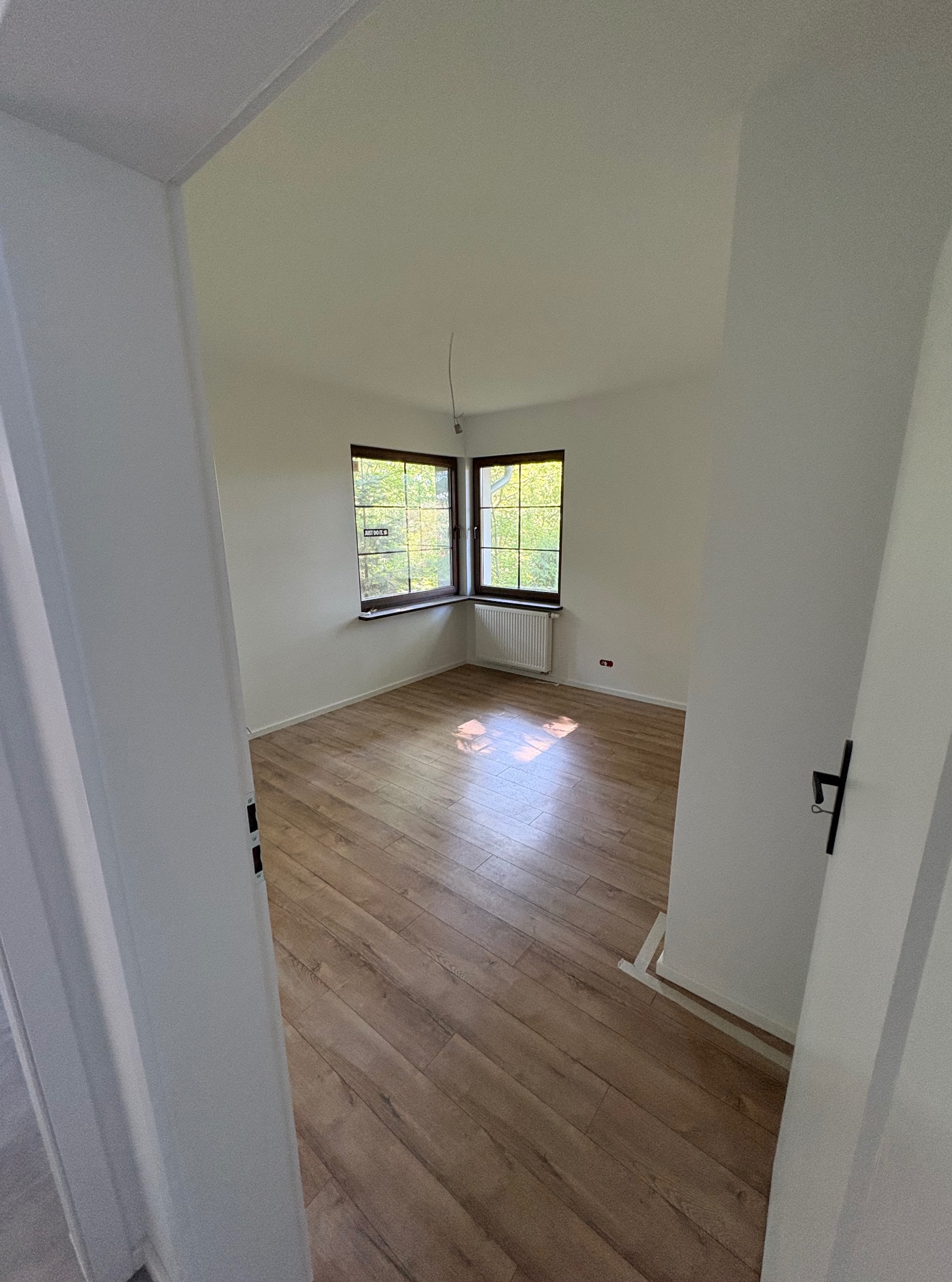 Pusty pokój z drewnianą podłogą, widok zza otwartych drzwi. Dwa okna z widokiem na zieleń. Świeżo pomalowane białe ściany. Grzejnik pod oknem.