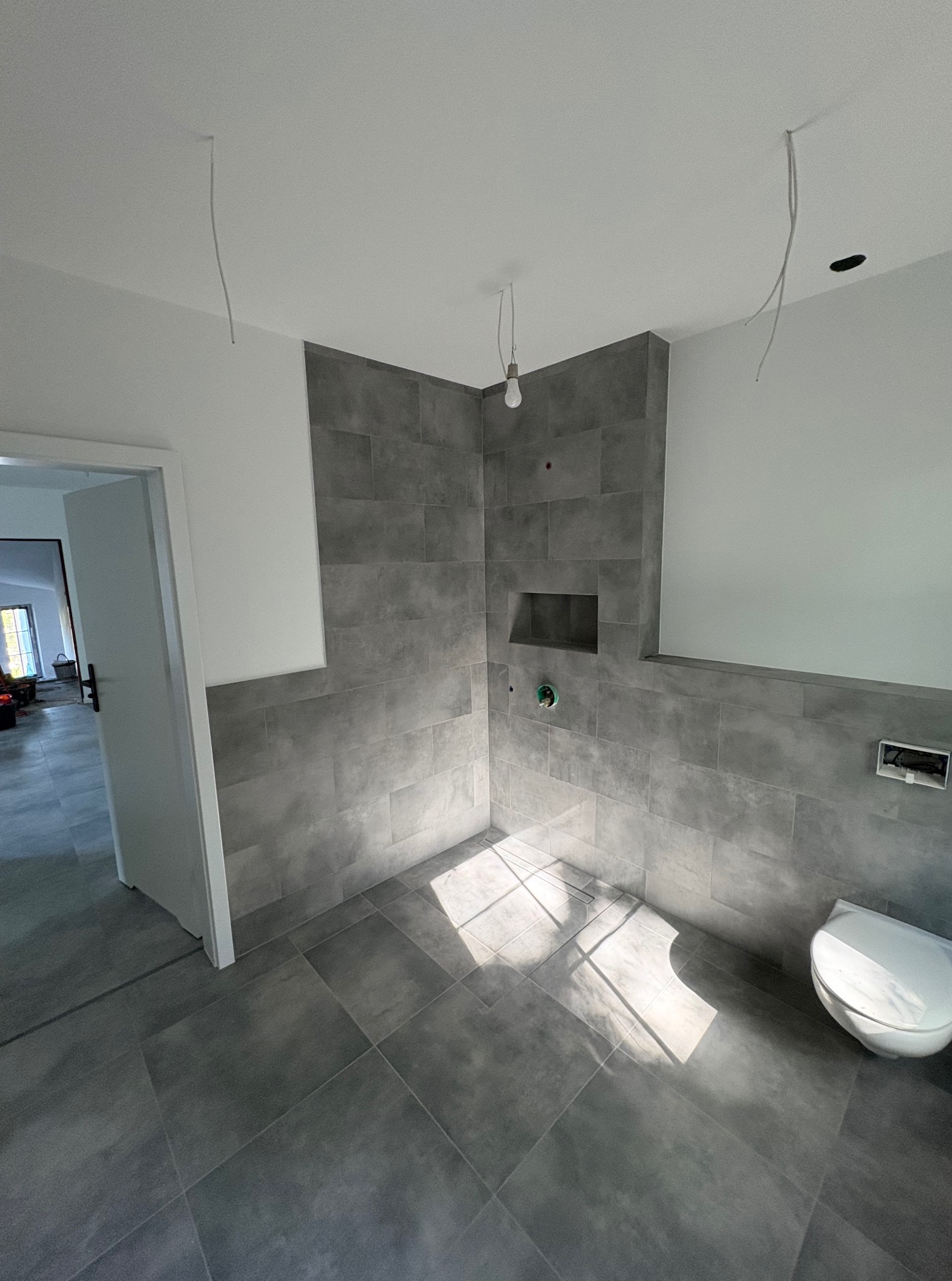 Łazienka w trakcie wykańczania: szare płytki, białe ściany, surowa żarówka. Widoczne niedokończone instalacje elektryczne i sanitarna. W tle otwarte drzwi.