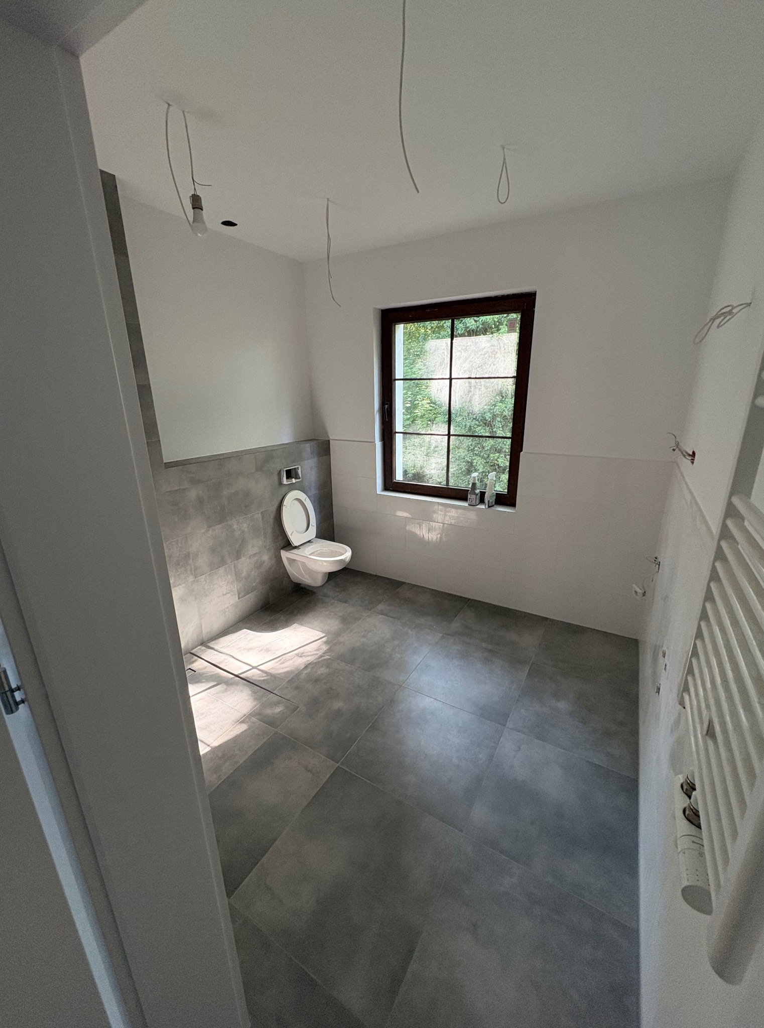 Wykończona łazienka w szaro-białej tonacji z oknem. Widoczne płytki na podłodze i ścianach, toaleta, grzejnik drabinkowy oraz wystające kable.