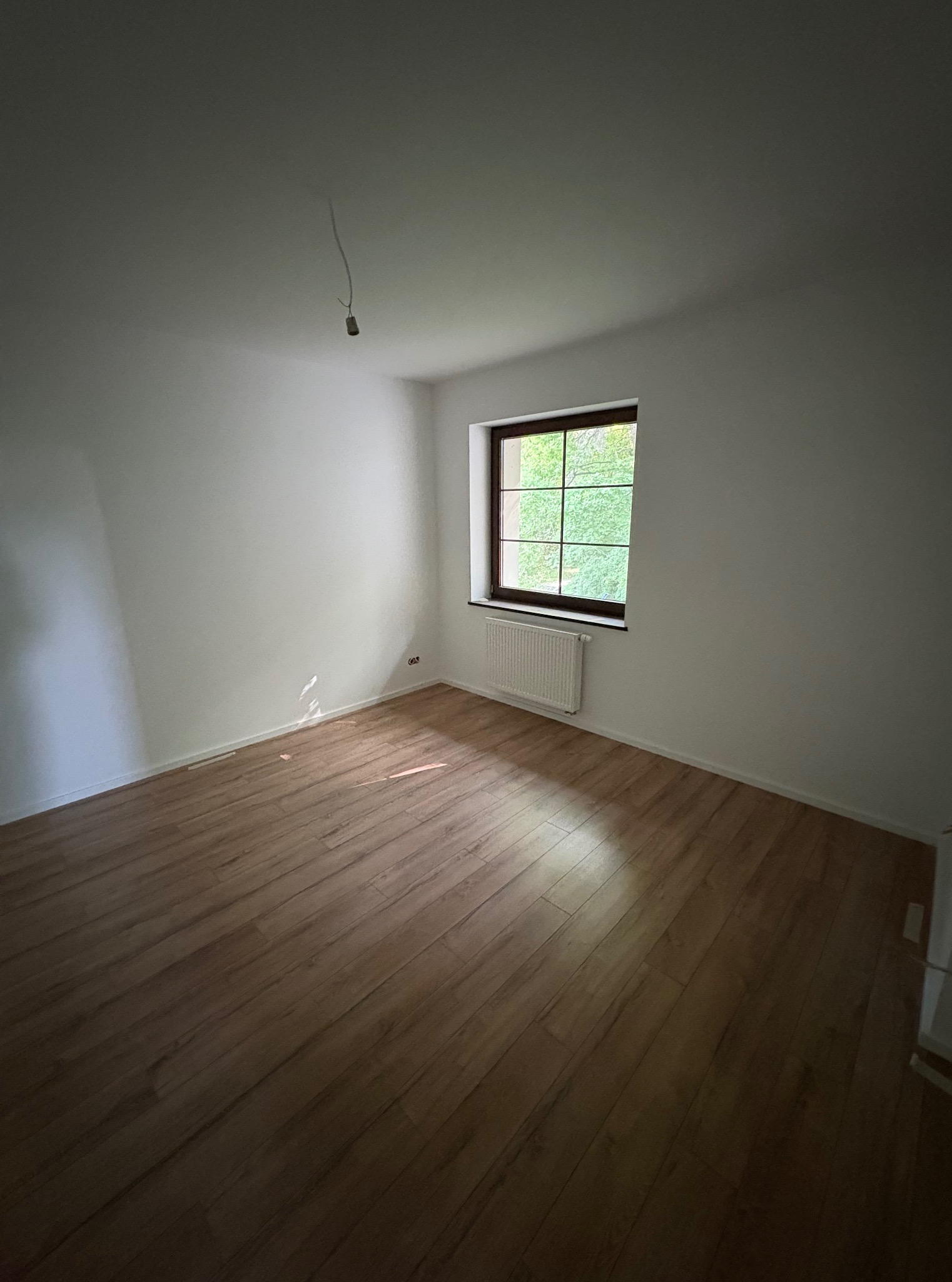 Pusty pokój z drewnianą podłogą, białymi ścianami i oknem z widokiem na zieleń. Na suficie widoczny kabel. Świeżo wykończone wnętrze.