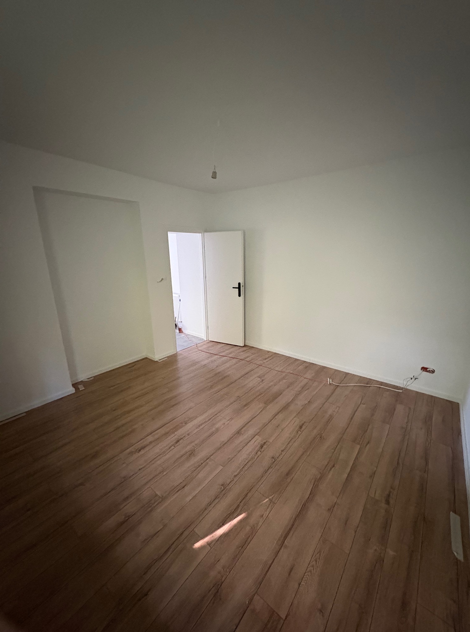 Puste, jasne pomieszczenie z panelami podłogowymi w odcieniach brązu. Widoczne białe ściany, otwarte drzwi i wystające kable elektryczne przy podłodze.