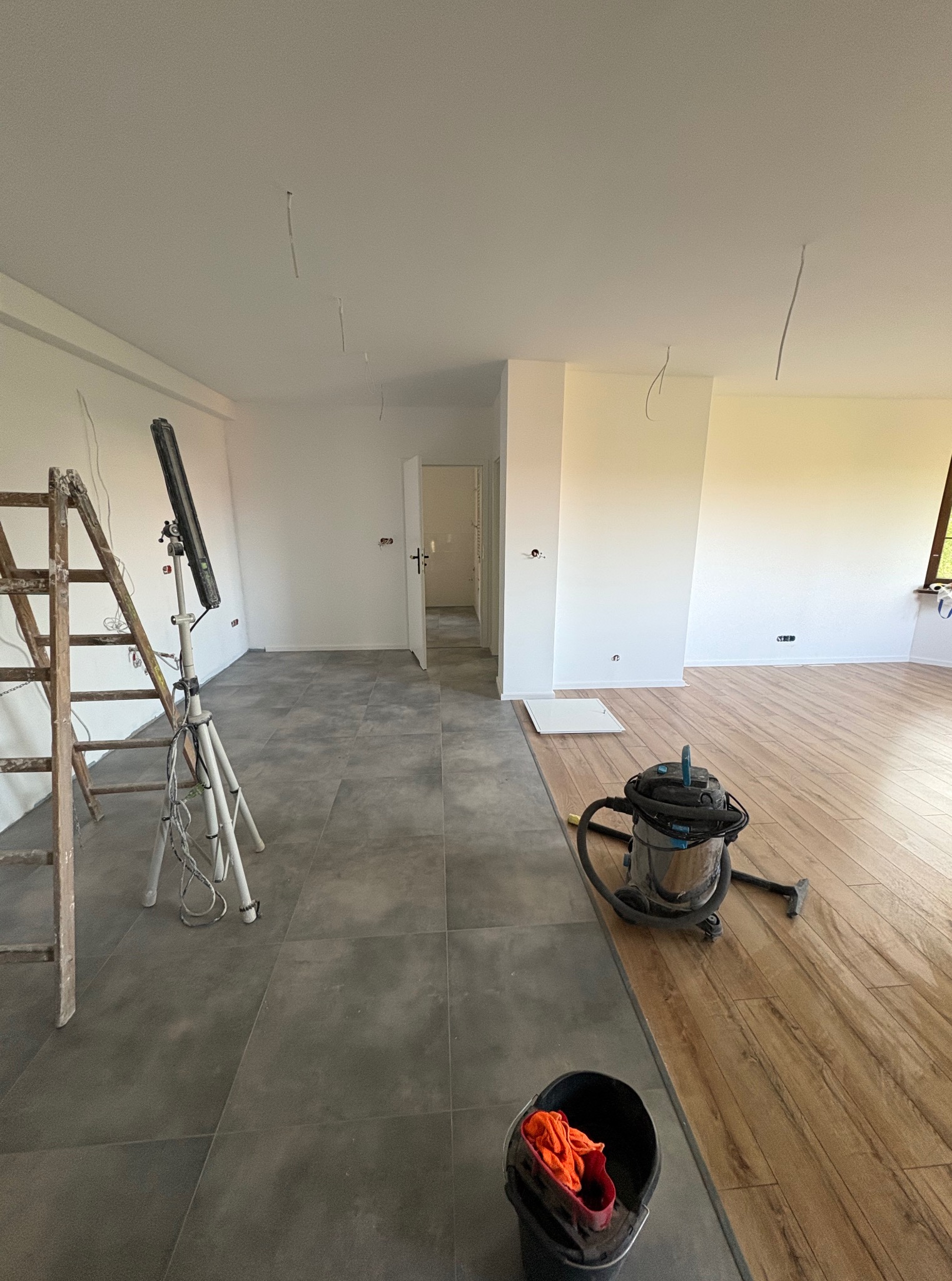 Wnętrze w trakcie prac wykończeniowych: ułożona podłoga (płytki i panele), drabina, lampa budowlana, odkurzacz i wiadro. Widoczne kable wystające ze ściany.
