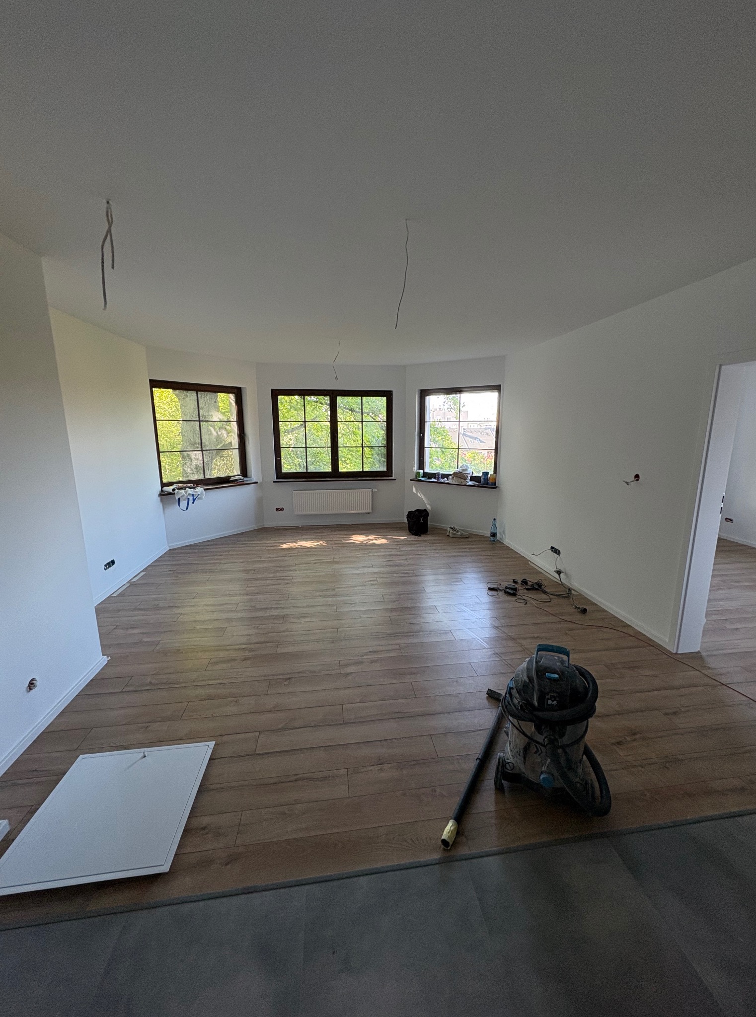 Jasne wnętrze po remoncie z drewnianą podłogą, trzema oknami i sprzętem budowlanym. Widoczne kable zwisające z sufitu i listwa przypodłogowa.