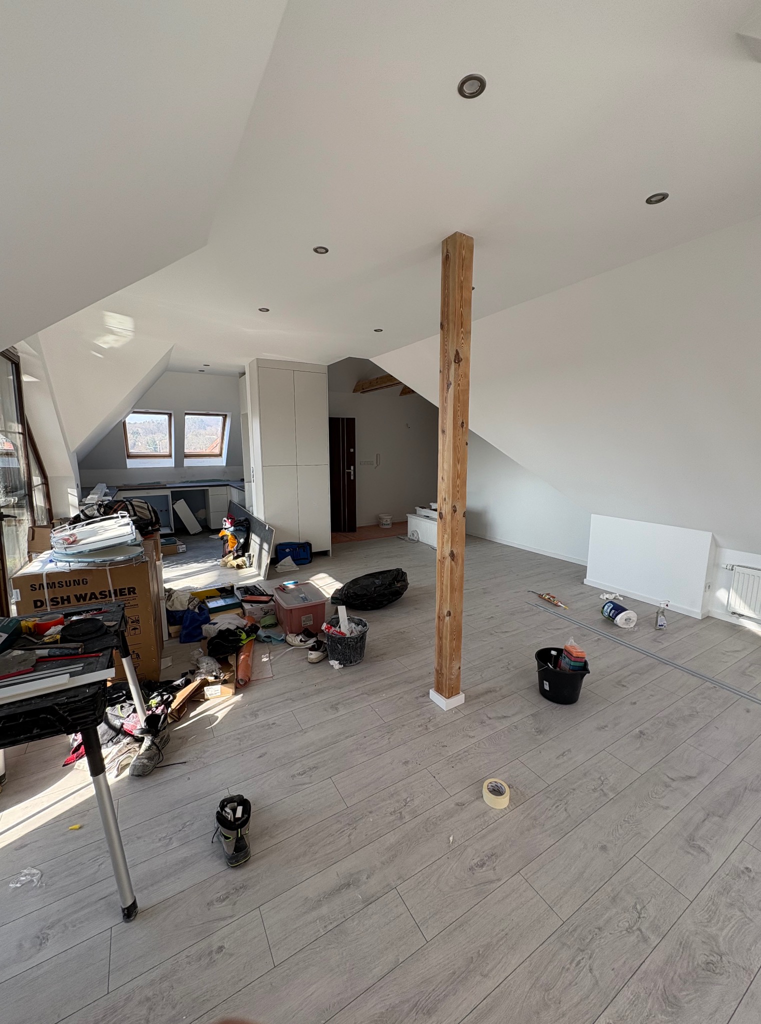 Poddasze w trakcie prac wykończeniowych: drewniany słup, szare panele, białe ściany i sufit, okna dachowe, widoczne narzędzia i materiały budowlane.