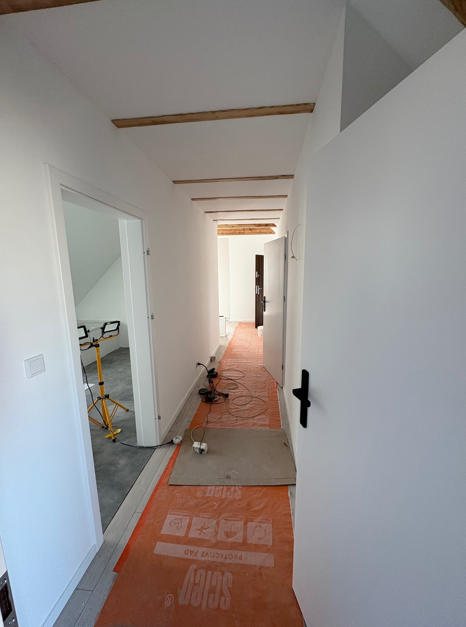 Korytarz w trakcie remontu: zabezpieczona podłoga pomarańczową folią, widoczne narzędzia, drewniane belki stropowe i świeżo pomalowane białe ściany. W tle otwarte drzwi.