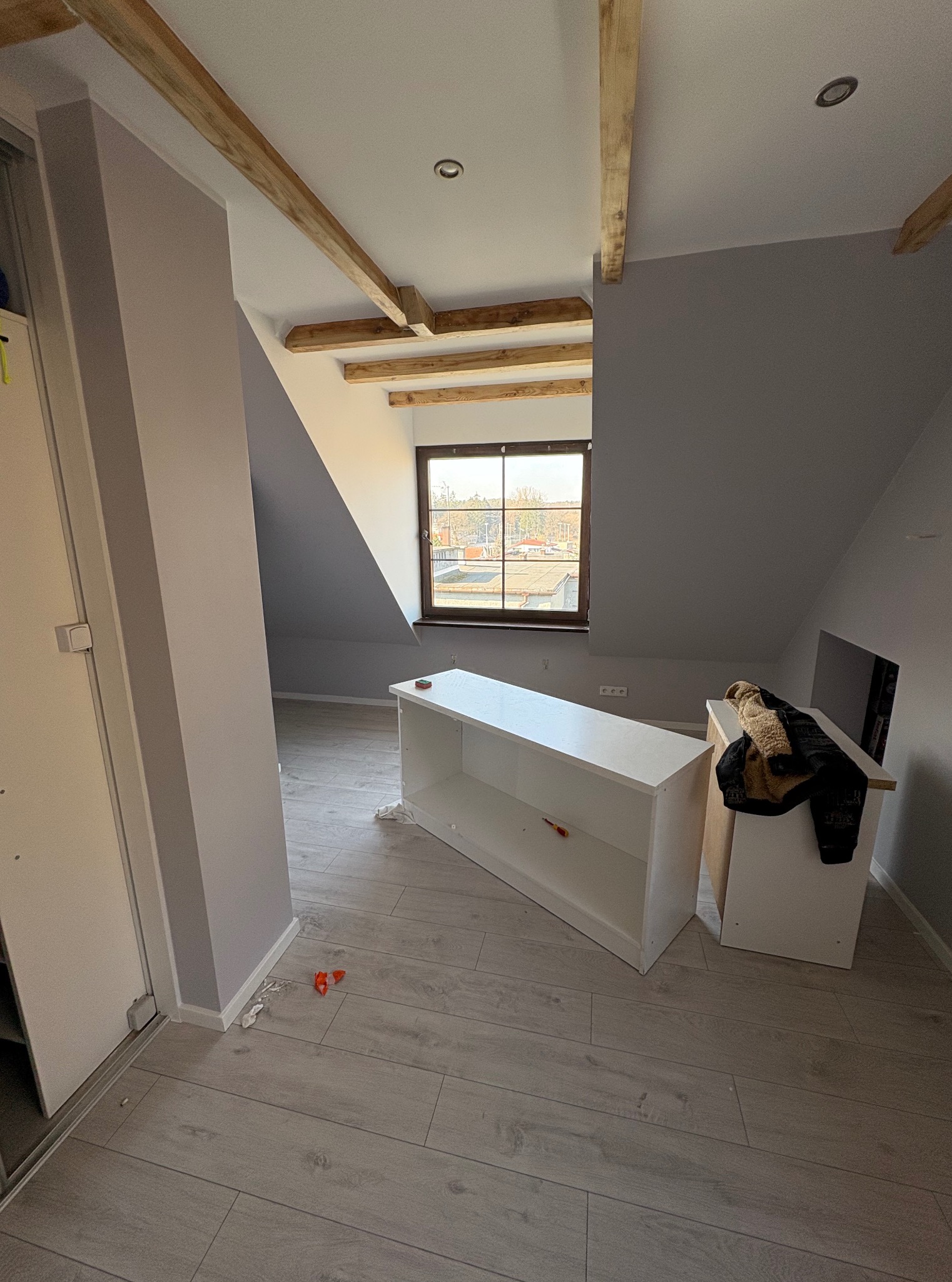 Poddasze z drewnianymi belkami stropowymi, jasnymi ścianami i oknem. Na podłodze panele i dwa białe regały przed montażem. Widok na zewnątrz przez okno.