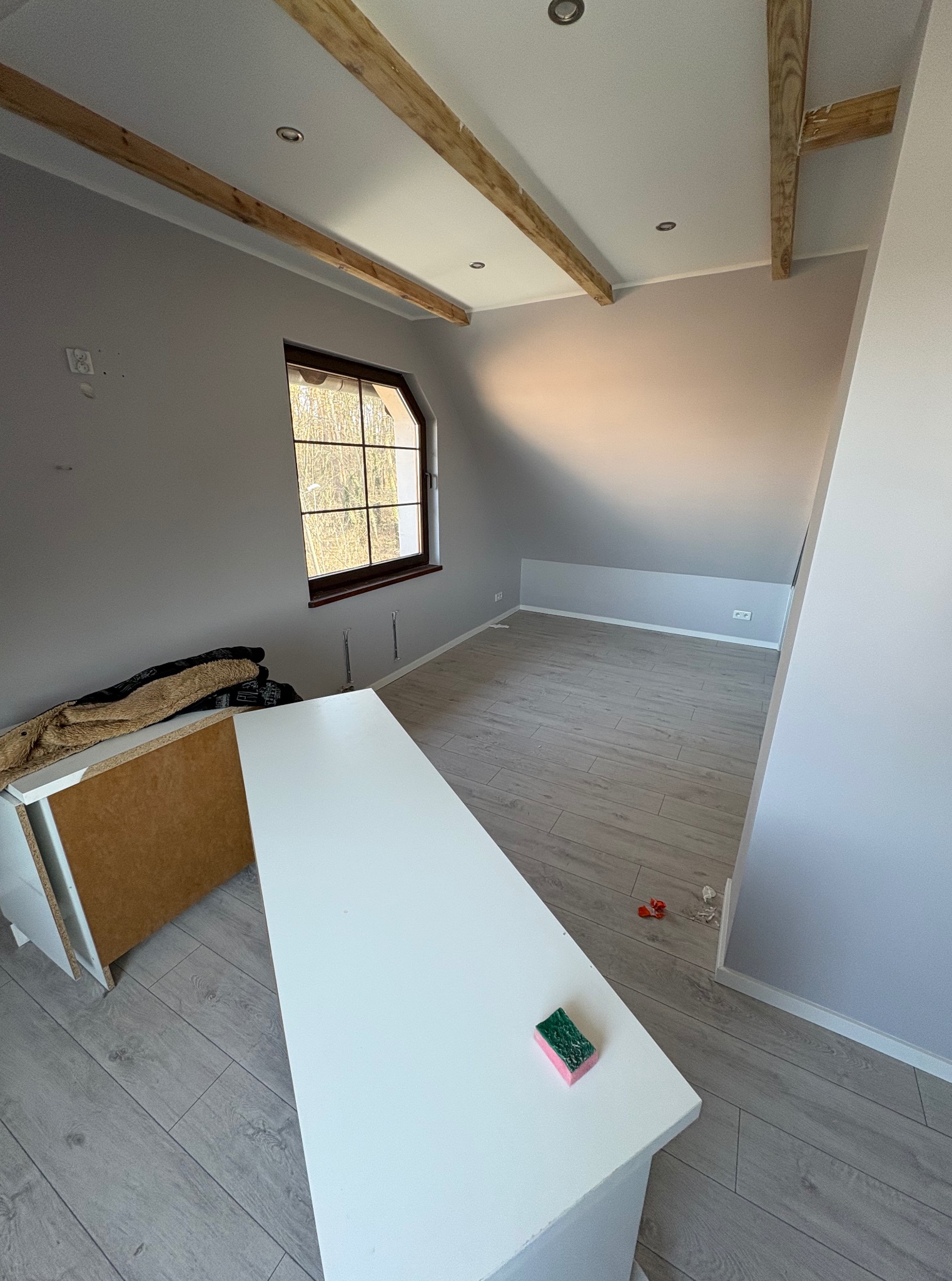 Szare wnętrze poddasza z drewnianymi belkami stropowymi, oknem i panelami podłogowymi. Widoczny fragment mebla i gąbka na blacie. Ściany pomalowane na szaro.