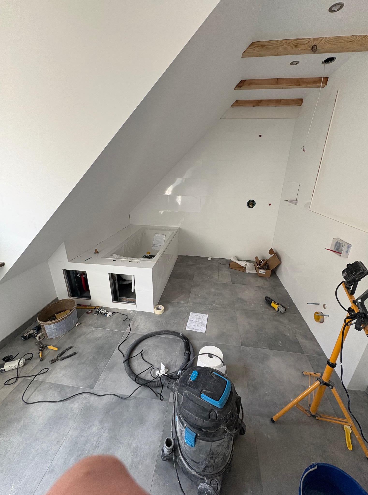 Remont łazienki na poddaszu: biała obudowa wanny w zabudowie, szare płytki, narzędzia i lampa budowlana. Widoczne elementy instalacji pod wanną.