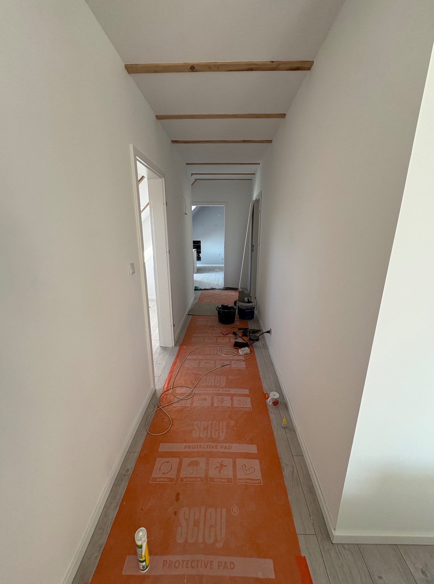 Korytarz w trakcie prac wykończeniowych: świeżo pomalowane ściany, zabezpieczona podłoga pomarańczową folią ochronną, widoczne drewniane elementy pod sufitem.