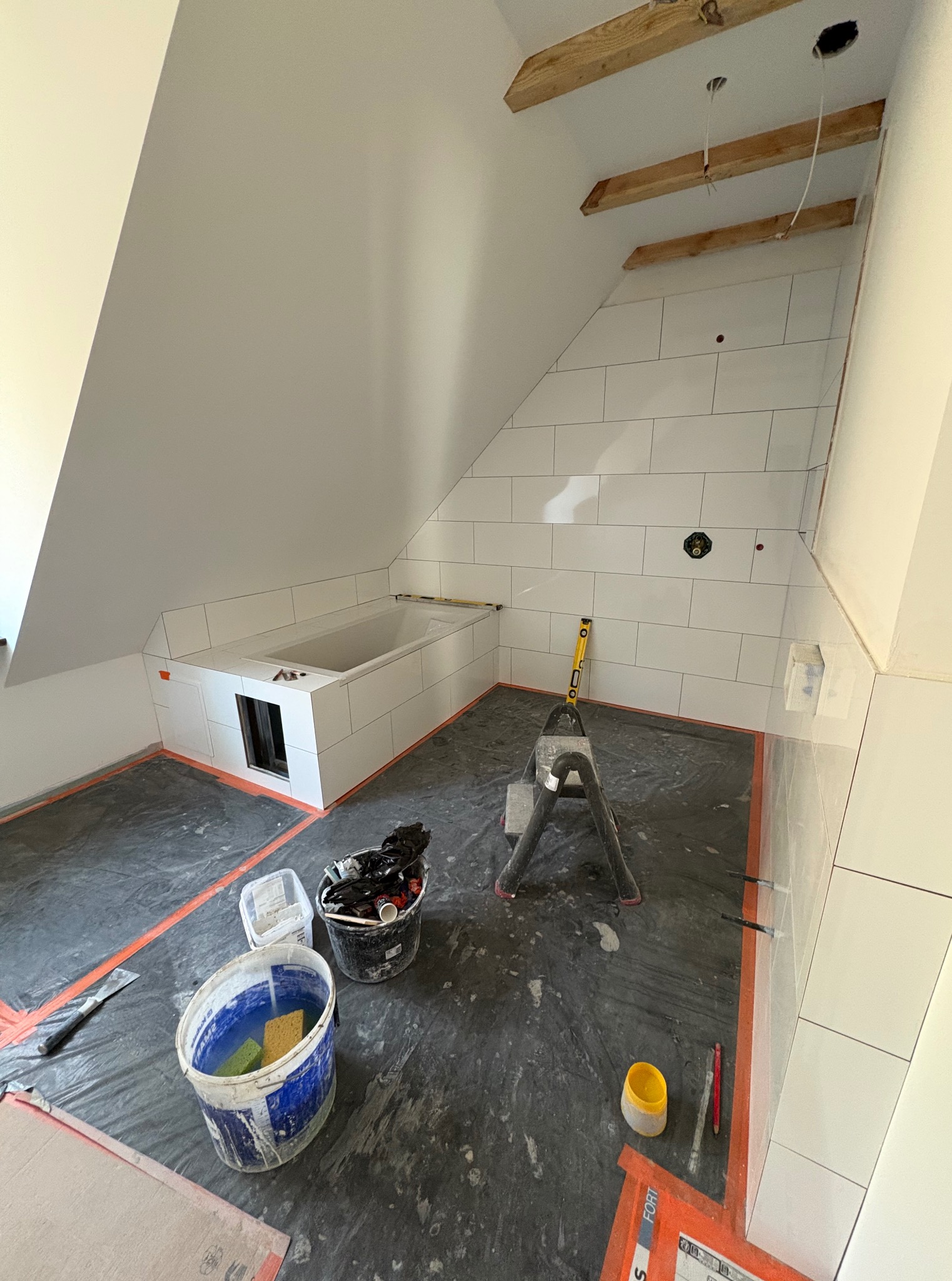 Białe płytki ułożone w łazience na poddaszu, wanna obudowana płytkami, drabina i narzędzia glazurnicze widoczne na folii ochronnej.