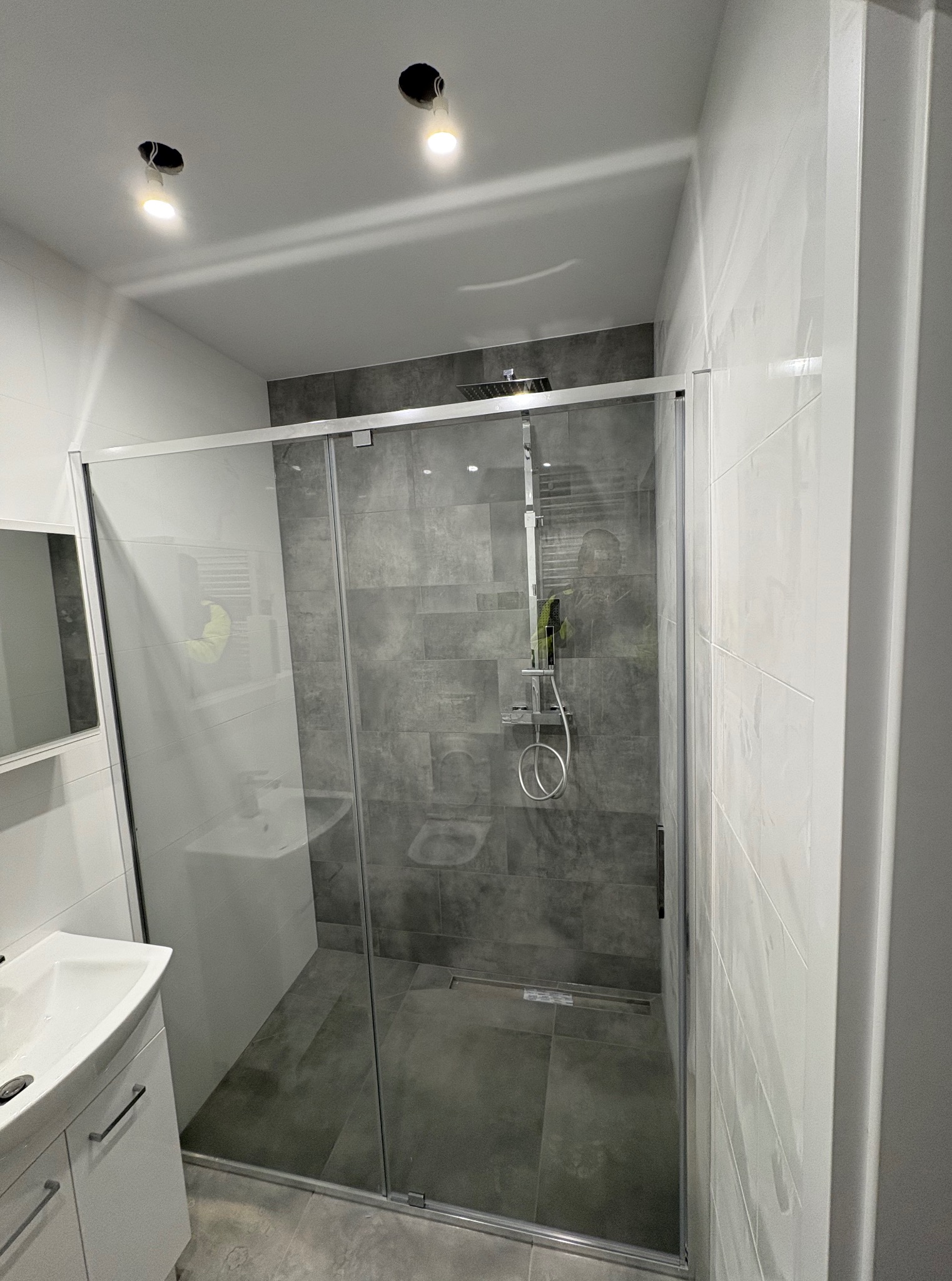 Nowoczesna kabina prysznicowa z szarymi płytkami, biała umywalka z szafką, oświetlenie sufitowe. Minimalistyczny design łazienki po remoncie.