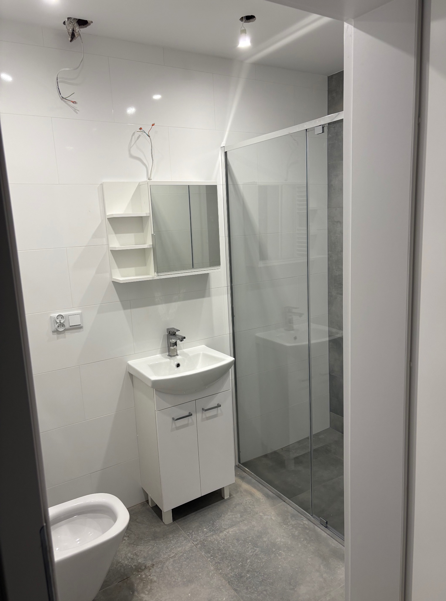 Nowoczesna łazienka z białą armaturą i szklaną kabiną prysznicową. Widoczne niedokończone prace elektryczne przy suficie.