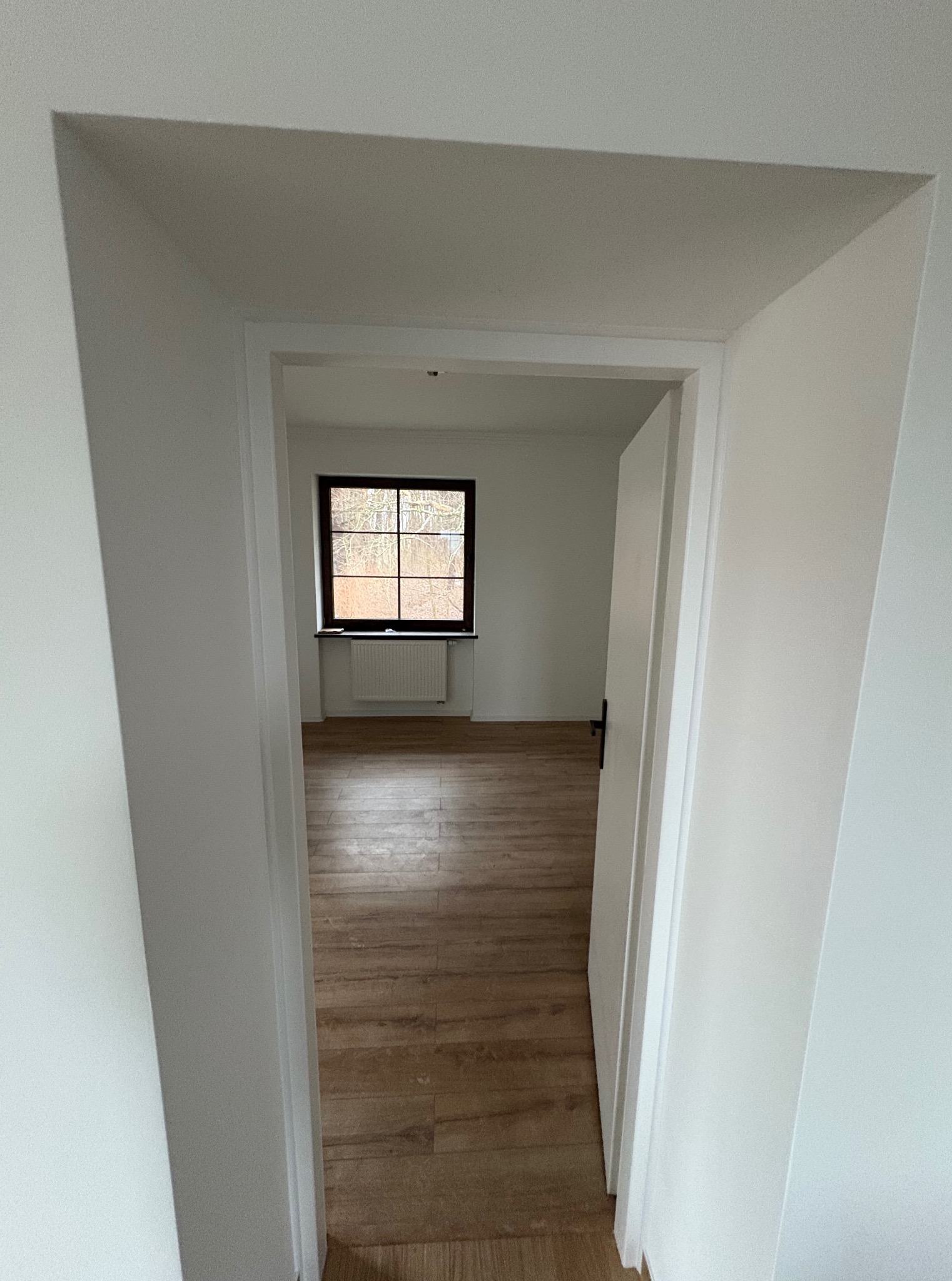 Widok przez otwarte drzwi na pokój z drewnianą podłogą i oknem. Świeżo pomalowane białe ściany i grzejnik pod oknem. Minimalistyczne wykończenie.