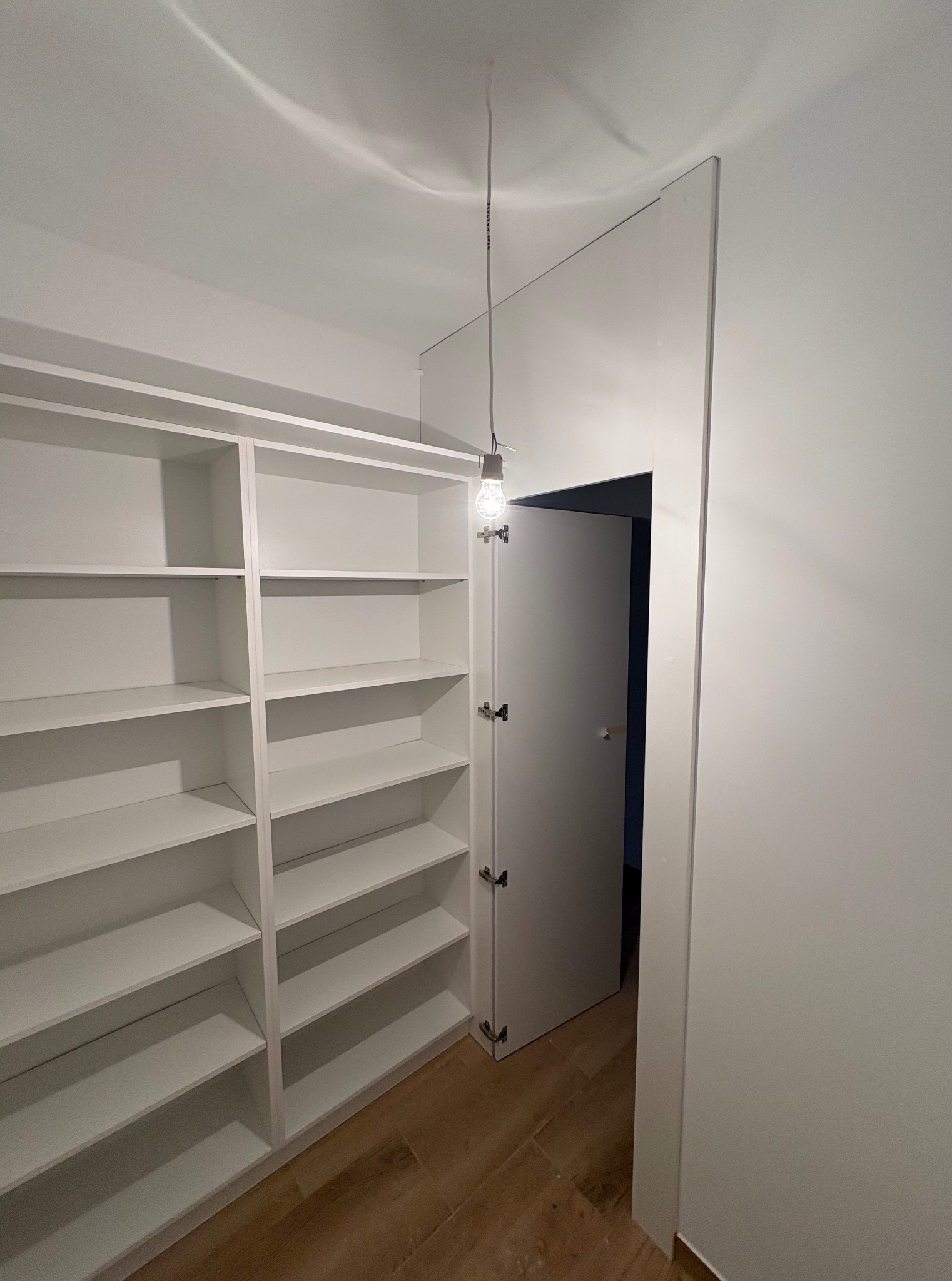 Biała zabudowa szafy wnękowej z półkami, widok z wnętrza pomieszczenia, z otwartymi drzwiami i żarówką. Minimalistyczny design, drewniana podłoga.