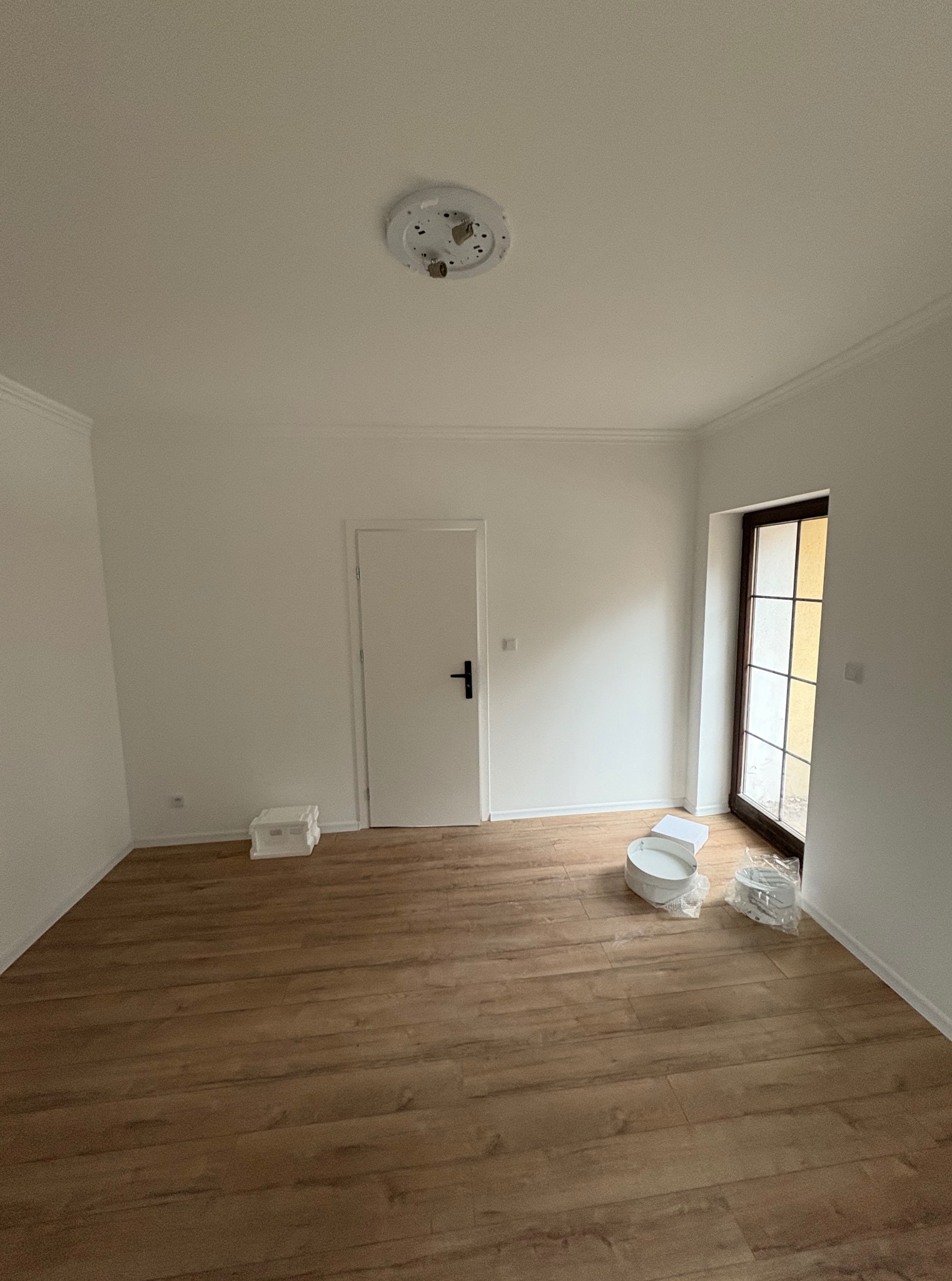 Pusty pokój po remoncie z drewnianą podłogą, białymi ścianami z listwami i drzwiami. Na podłodze widoczne akcesoria malarskie.