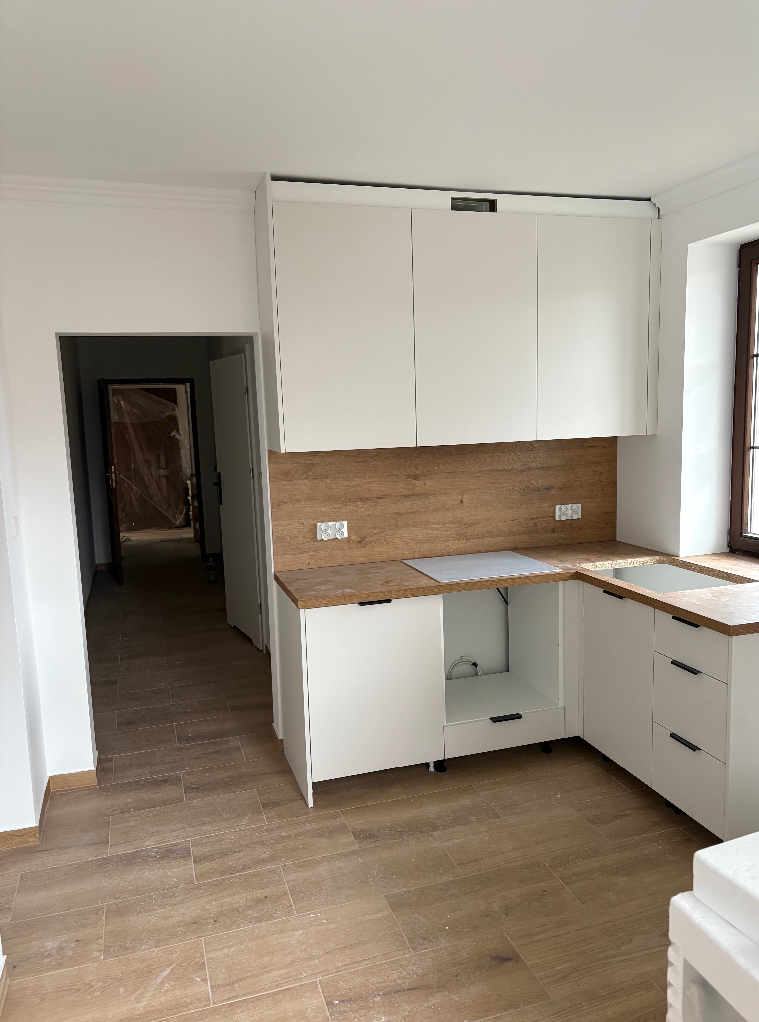 Jasna kuchnia w trakcie montażu: białe szafki z drewnianymi blatami i panelami, brązowe płytki na podłodze, widoczne gniazdka elektryczne i otwarte przejście do innego pomieszczenia.