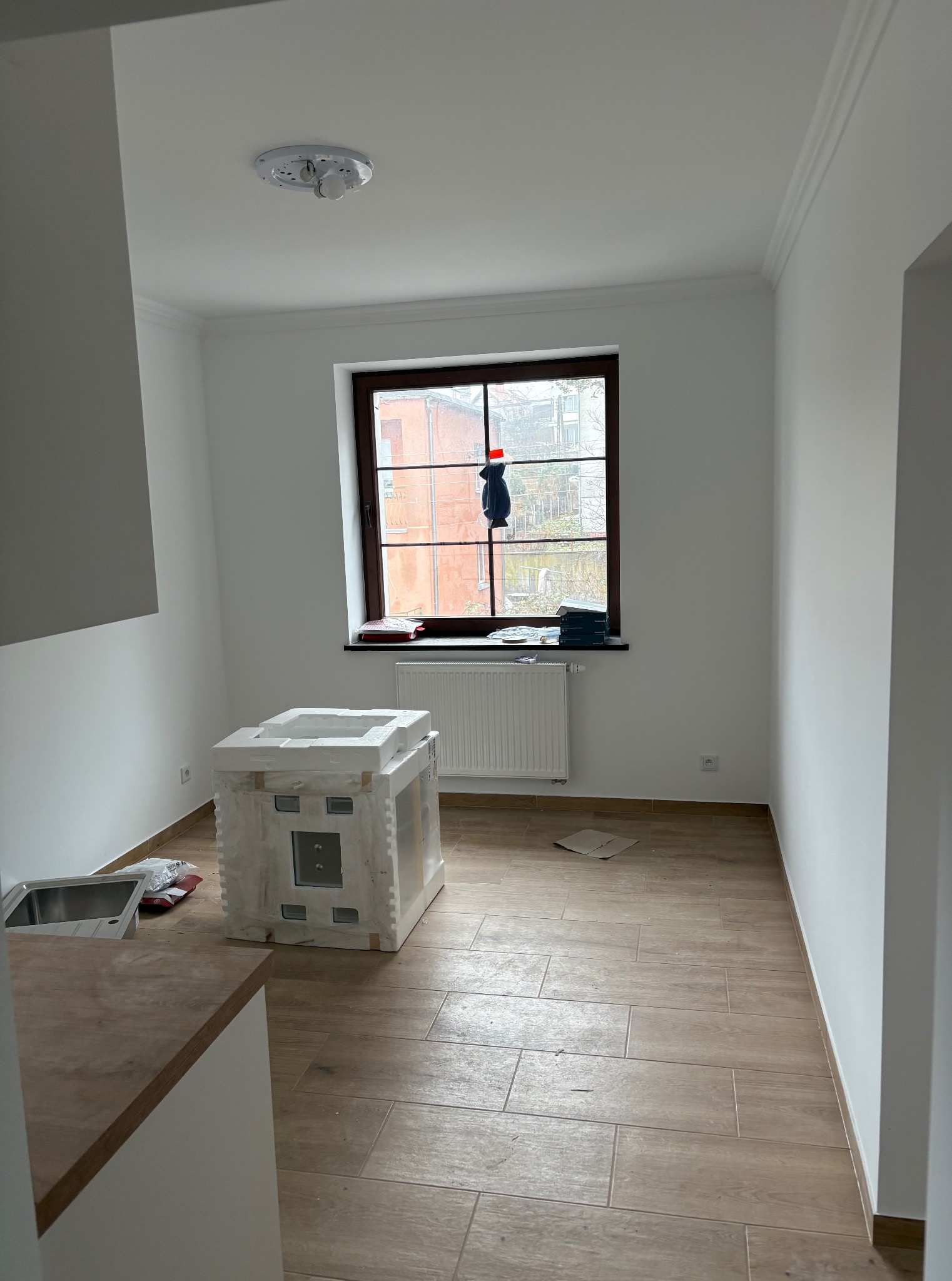 Jasne wnętrze z drewnopodobną podłogą. Okno z brązową ramą, białe ściany z listwami. W pomieszczeniu znajduje się zlew, grzejnik i zapakowany sprzęt.