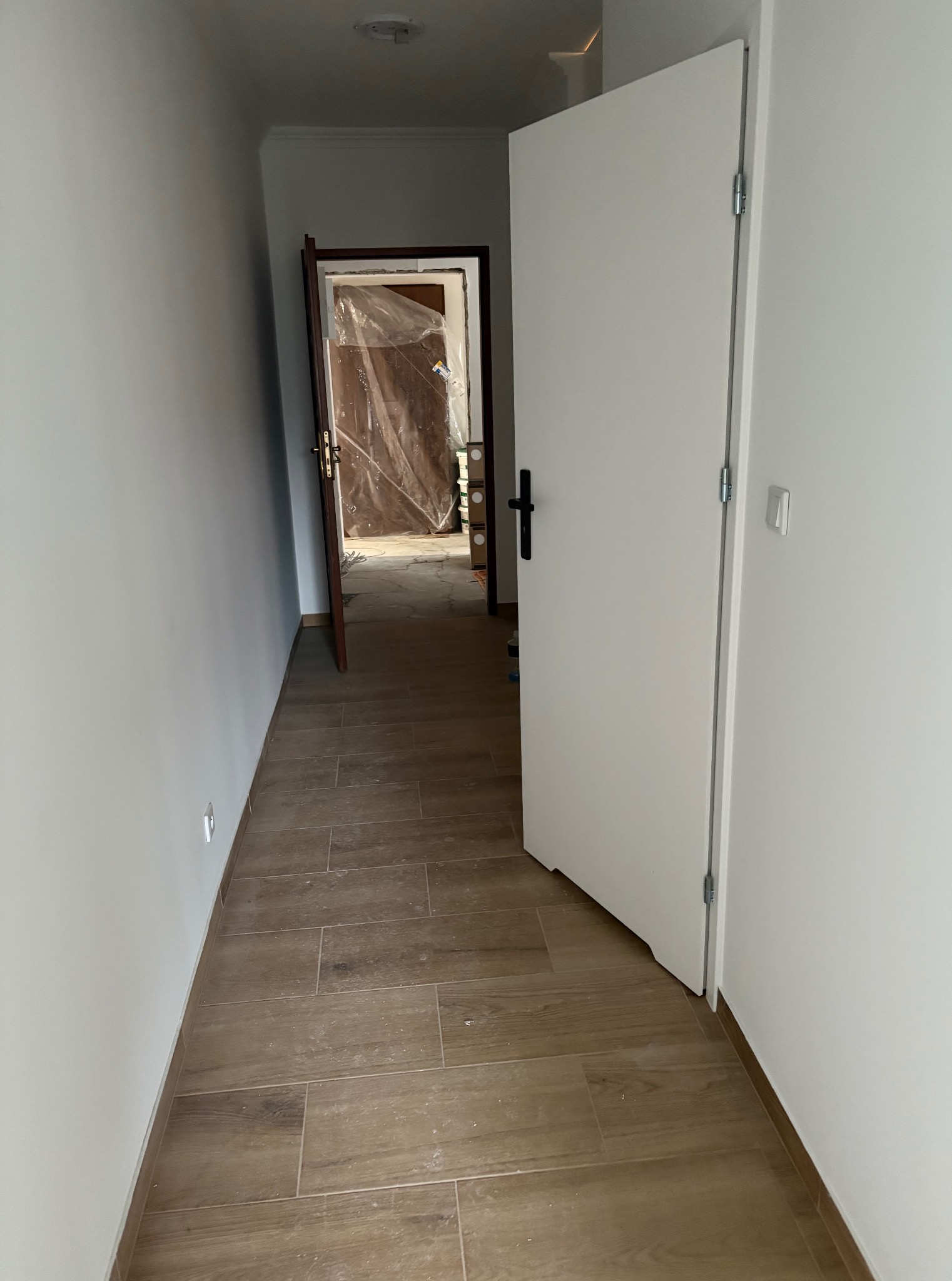 Korytarz po remoncie: białe ściany, brązowe płytki podłogowe imitujące drewno, otwarte białe drzwi i widok na zakryte folią meble w kolejnym pomieszczeniu.