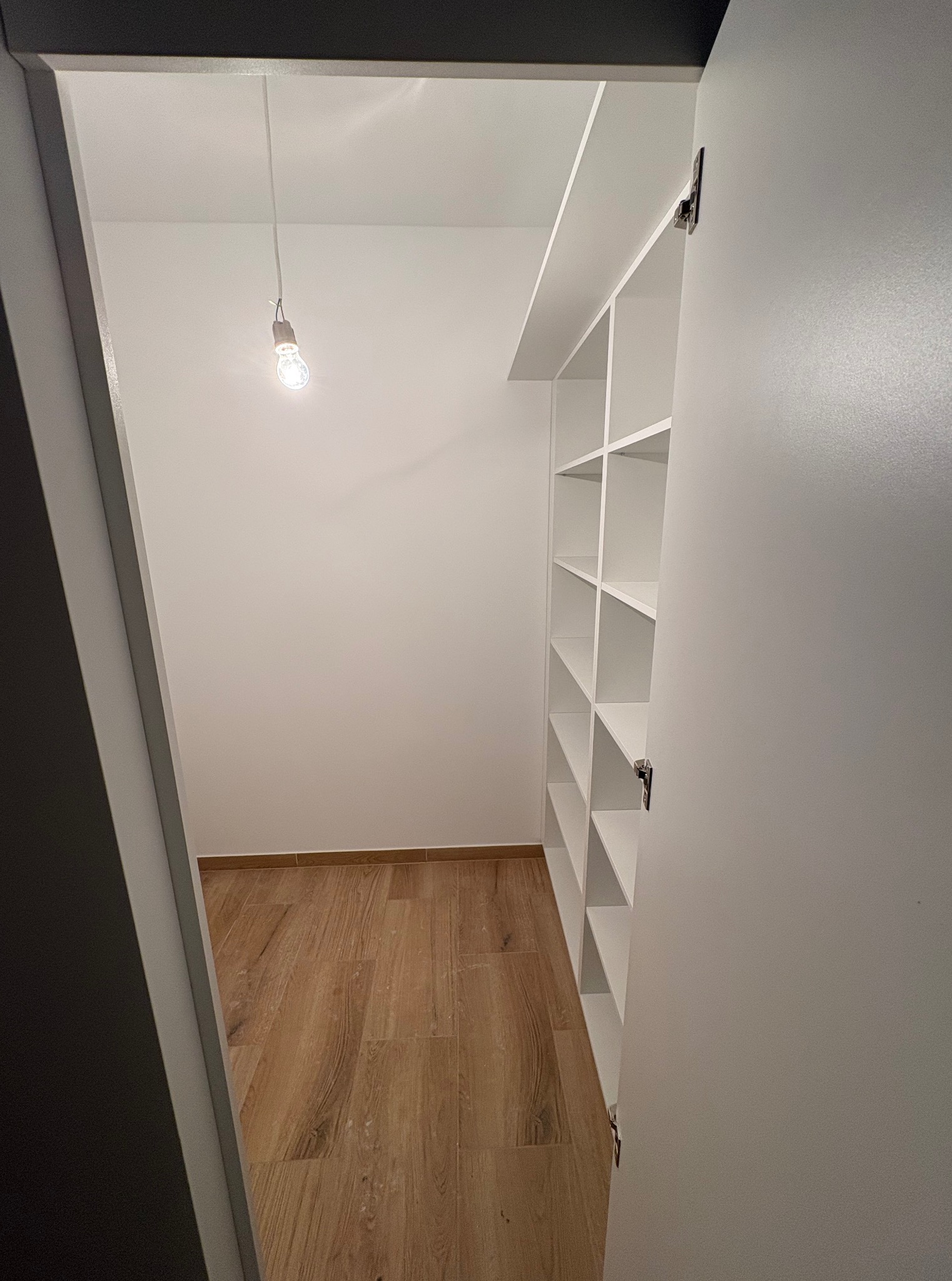 Wnętrze garderoby po remoncie: drewniana podłoga, białe ściany i regał z półkami. Widoczne oświetlenie w postaci wiszącej żarówki. Kątowe ujęcie z otwartymi drzwiami.