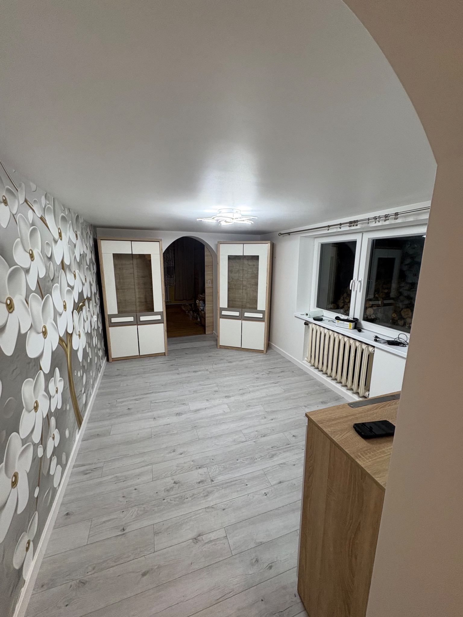 Wykończone wnętrze pokoju z szarą tapetą w kwiaty, drewnianą podłogą i białymi szafkami po bokach przejścia. Widoczny grzejnik pod oknem i fragment drewnianej komody.