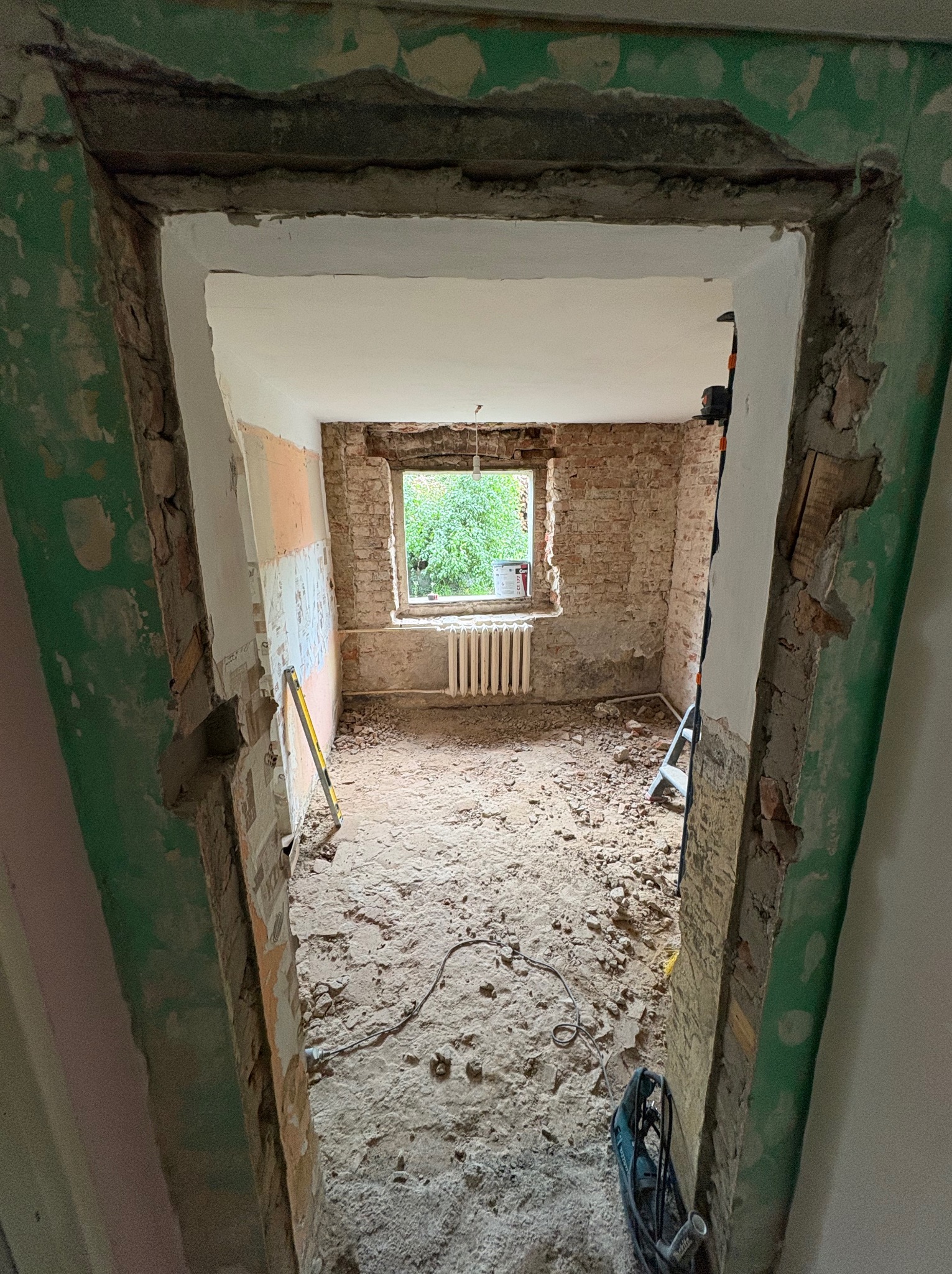 Pokój w trakcie demontażu, widok przez futrynę. Odsłonięta cegła, gruz na podłodze, poziomica oparta o ścianę. Widoczne narzędzia i drabina.