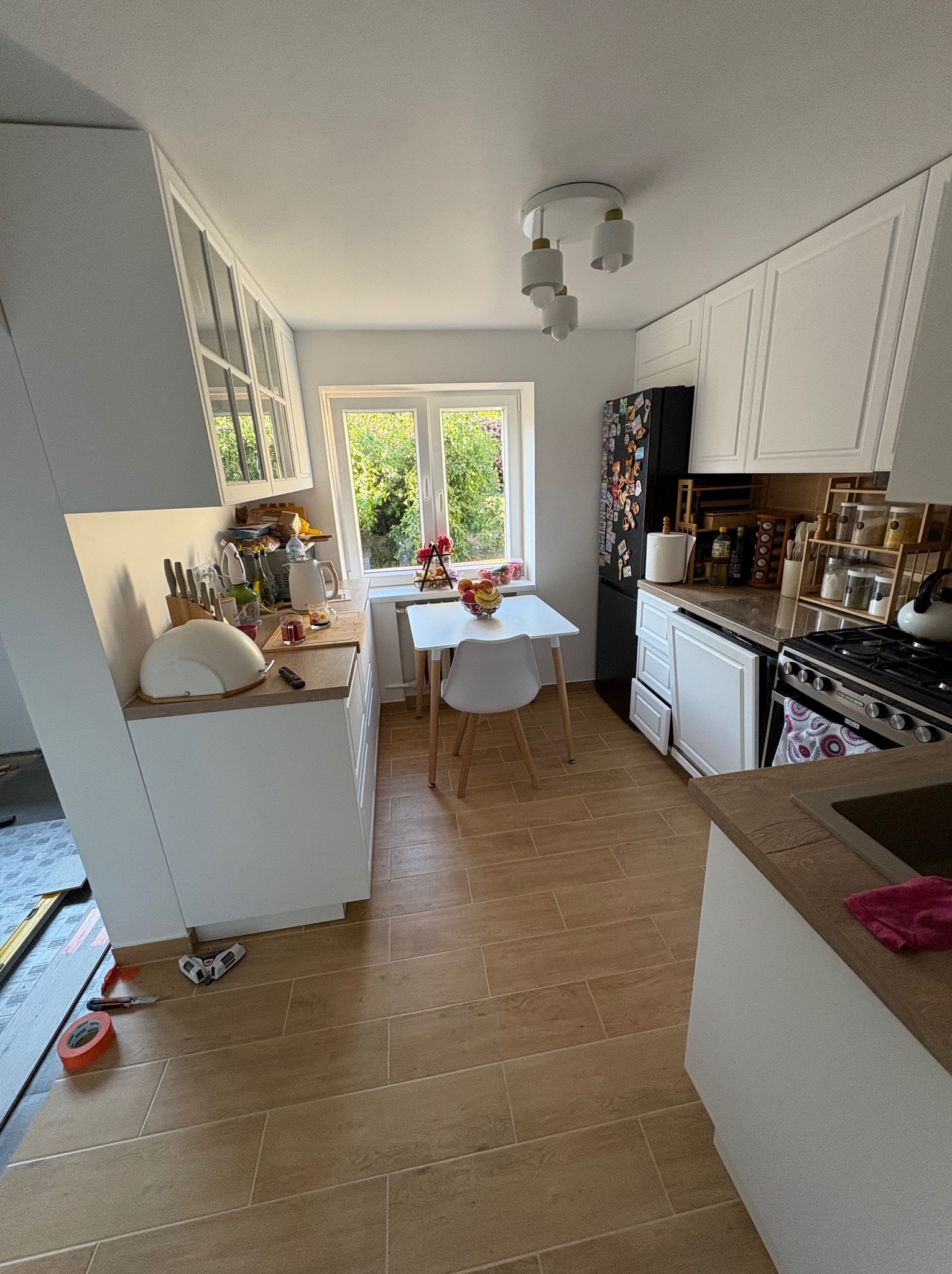 Jasna kuchnia po remoncie z drewnopodobną podłogą, białymi szafkami i stołem przy oknie. Widoczne narzędzia i materiały wskazują na trwające prace wykończeniowe.