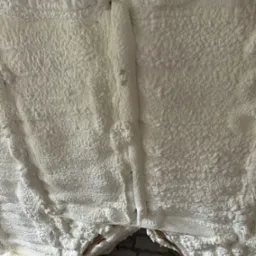 Szczegółowe ujęcie białej pianki PUR, gęsto wypełniającej przestrzeń poddasza, z widocznymi elementami konstrukcji drewnianej i fragmentami muru. Tekstura pianki nierównomierna.