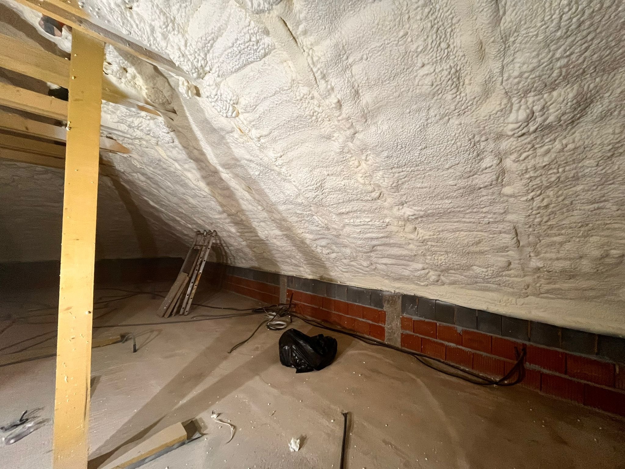 Poddasze po ociepleniu pianką PUR, widoczne krokwie i fragment muru. Izolacja o nieregularnej fakturze pokrywa skośne powierzchnie.