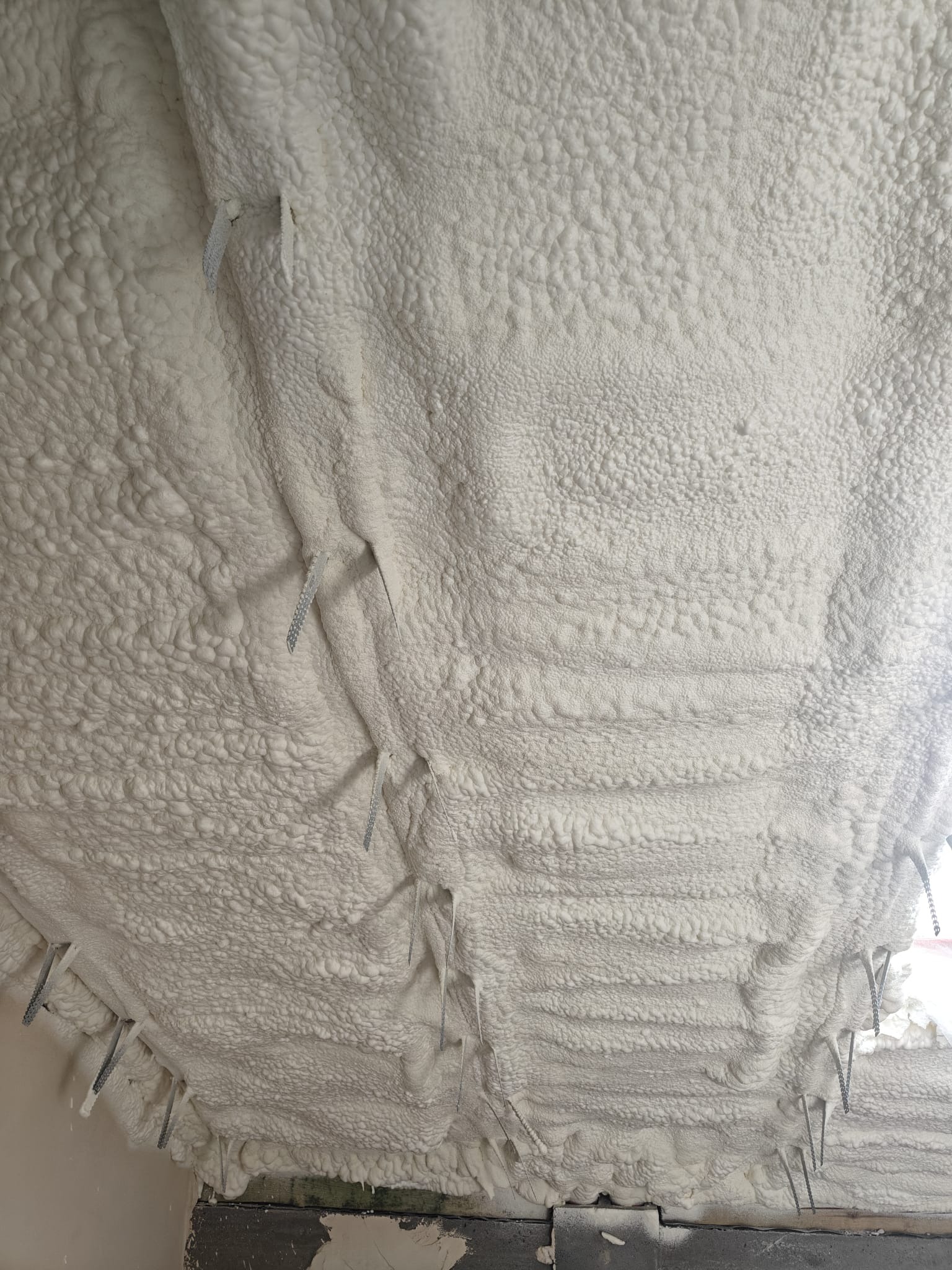 Poddasze po ociepleniu pianką PUR, widoczne metalowe elementy mocujące. Struktura piany gęsta, biała, z nierówną powierzchnią. Detal izolacji natryskowej.