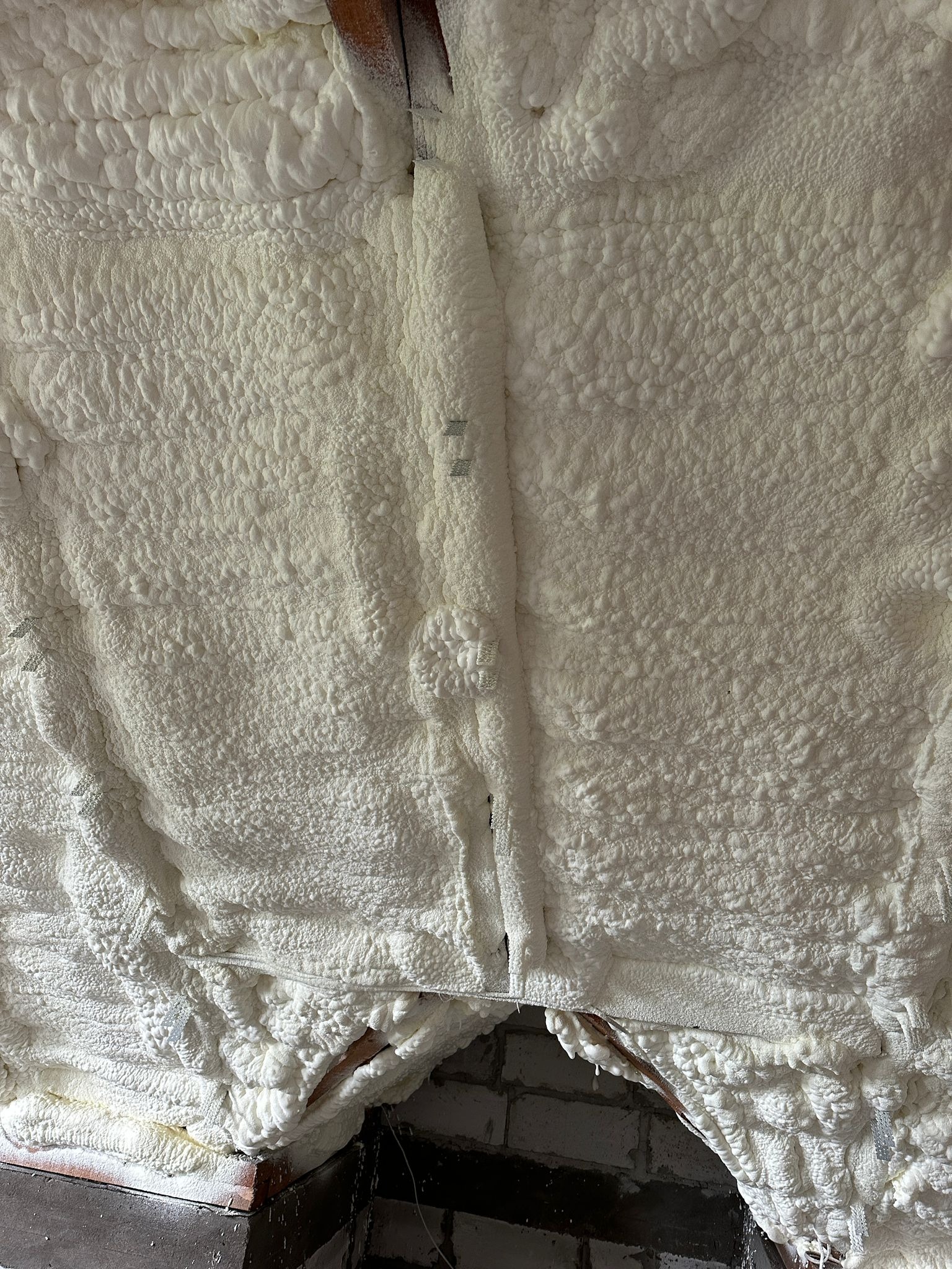 Szczegółowe ujęcie białej pianki PUR, gęsto wypełniającej przestrzeń poddasza, z widocznymi elementami konstrukcji drewnianej i fragmentami muru. Tekstura pianki nierównomierna.