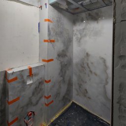 Flizenlega - Łazienka w trakcie wykańczania: marmurowe płytki na ścianach, zabezpieczone pomarańczową taśmą, widoczne elementy instalacji i surowy stan wykończenia.