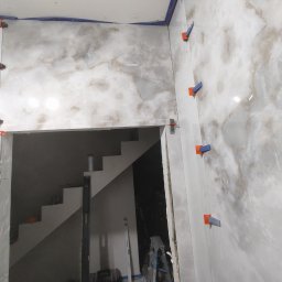 Flizenlega - Układanie płytek imitujących marmur na ścianie przy schodach, widoczne poziomice i system poziomowania płytek. Wnętrze w trakcie prac wykończeniowych.