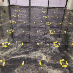 Flizenlega - Układanie płytek podłogowych z użyciem systemu poziomowania. Widoczne narzędzia glazurnicze, czarne płytki z marmurkowym wzorem i żółte kliny dystansowe.