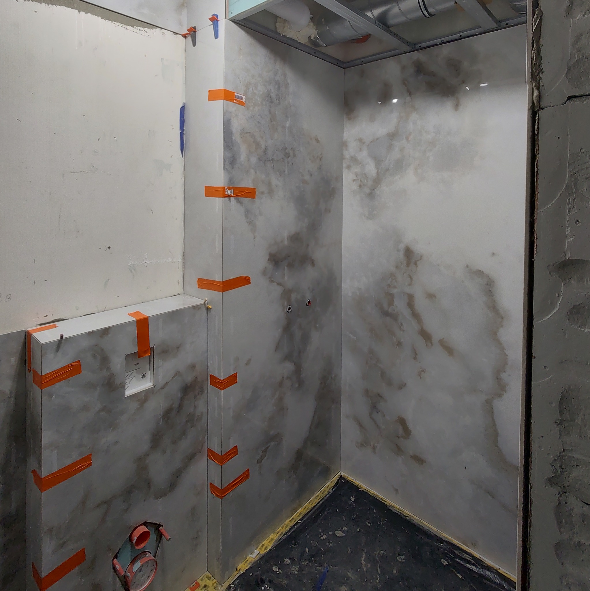 Łazienka w trakcie wykańczania: marmurowe płytki na ścianach, zabezpieczone pomarańczową taśmą, widoczne elementy instalacji i surowy stan wykończenia.