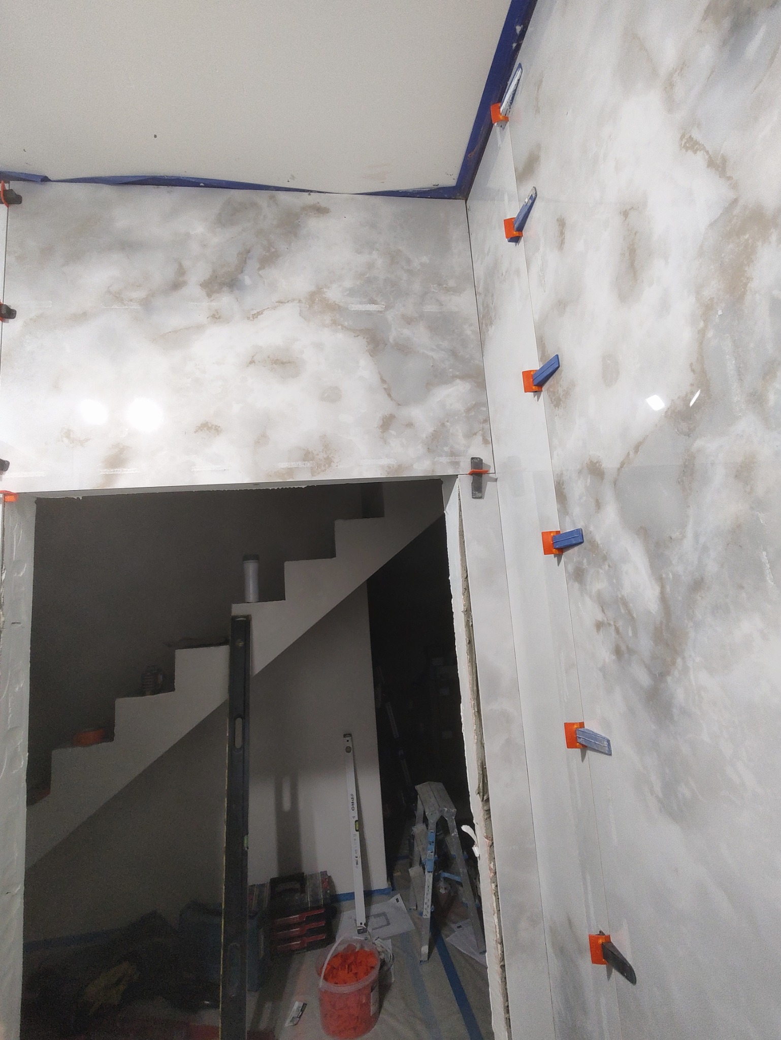 Układanie płytek imitujących marmur na ścianie przy schodach, widoczne poziomice i system poziomowania płytek. Wnętrze w trakcie prac wykończeniowych.