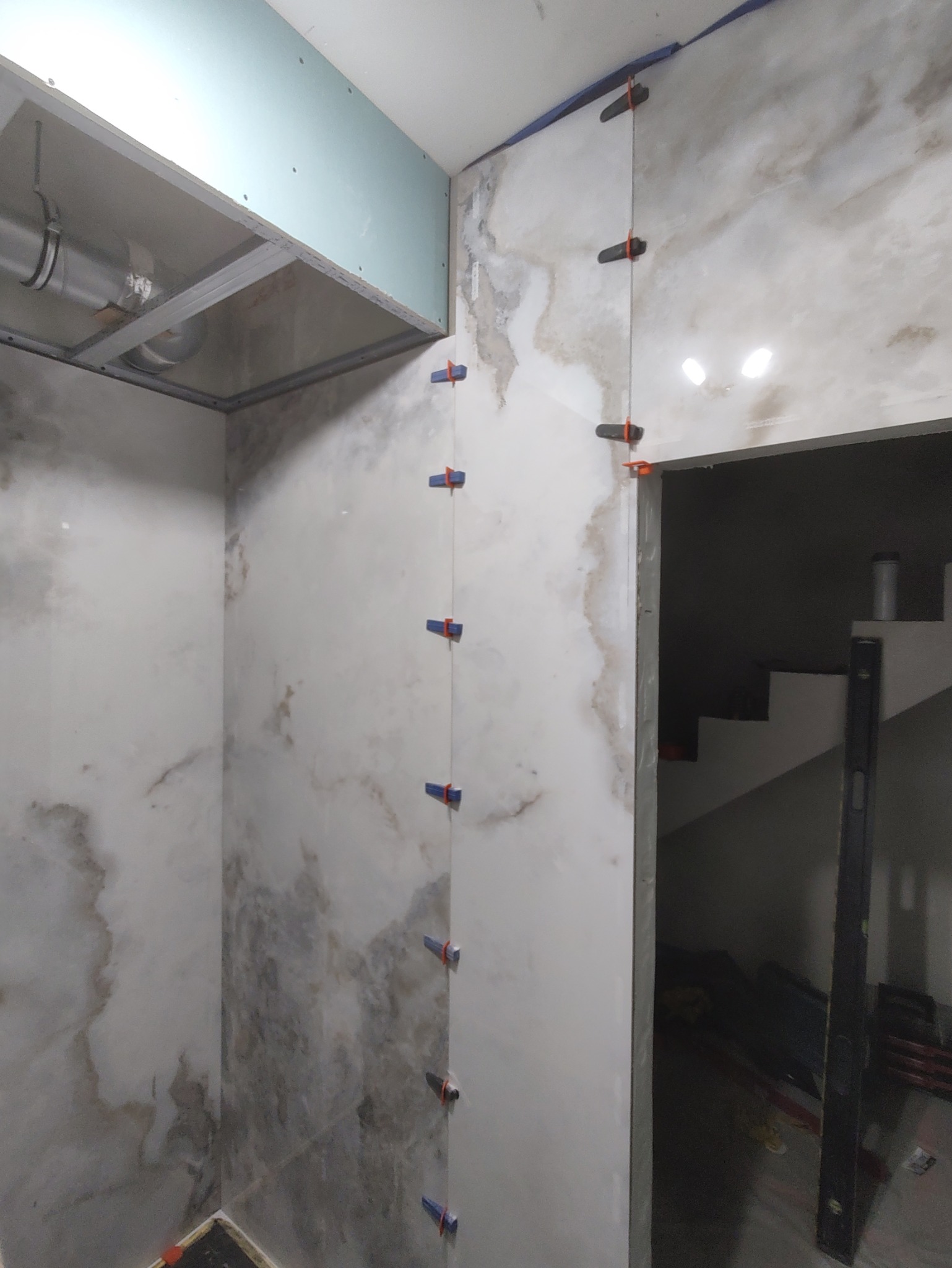 Ściany łazienki w trakcie układania płytek imitujących marmur, z widocznymi klipsami poziomującymi. Częściowo widoczna konstrukcja sufitu podwieszanego.