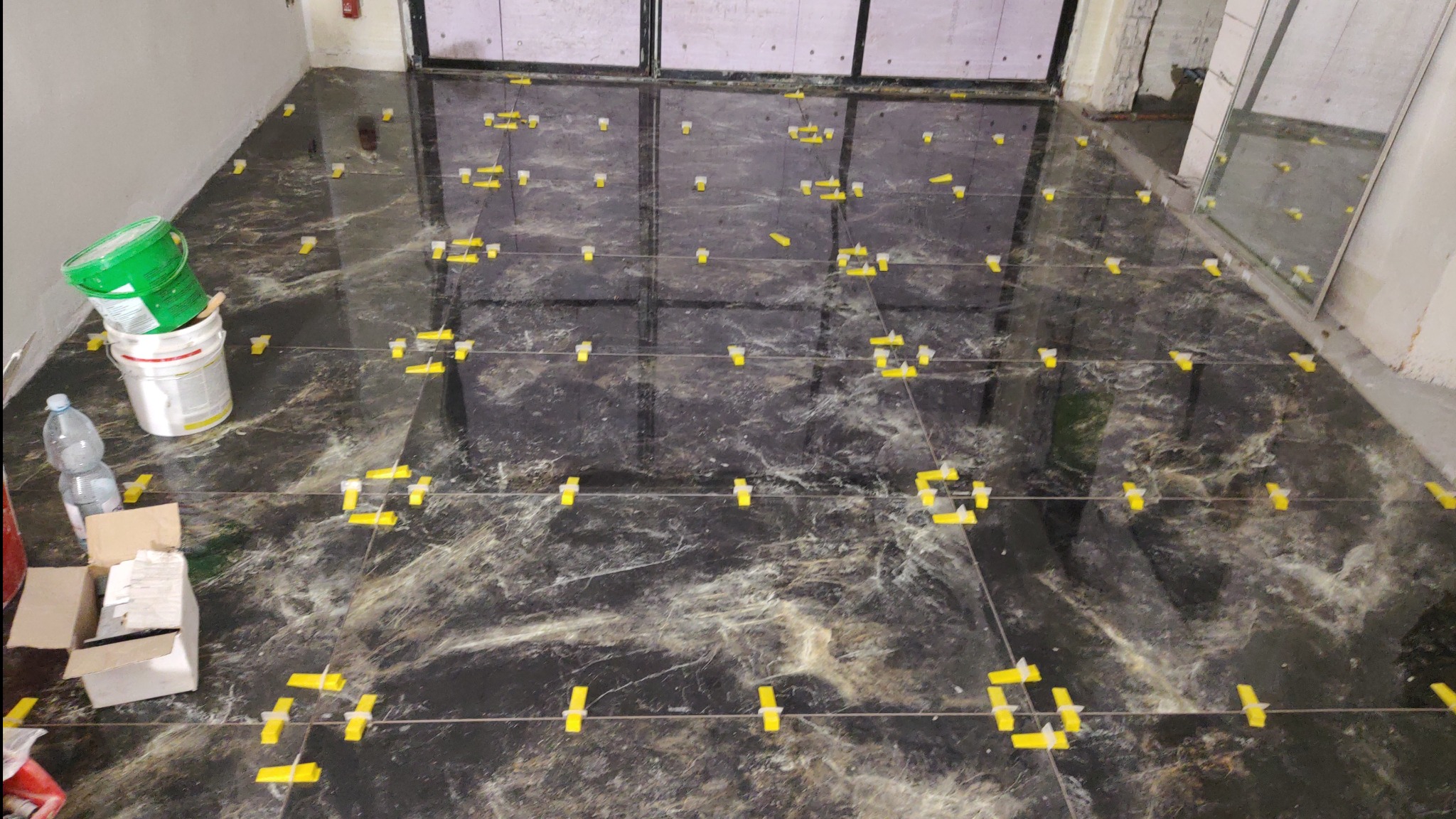 Układanie płytek podłogowych z użyciem systemu poziomowania. Widoczne narzędzia glazurnicze, czarne płytki z marmurkowym wzorem i żółte kliny dystansowe.