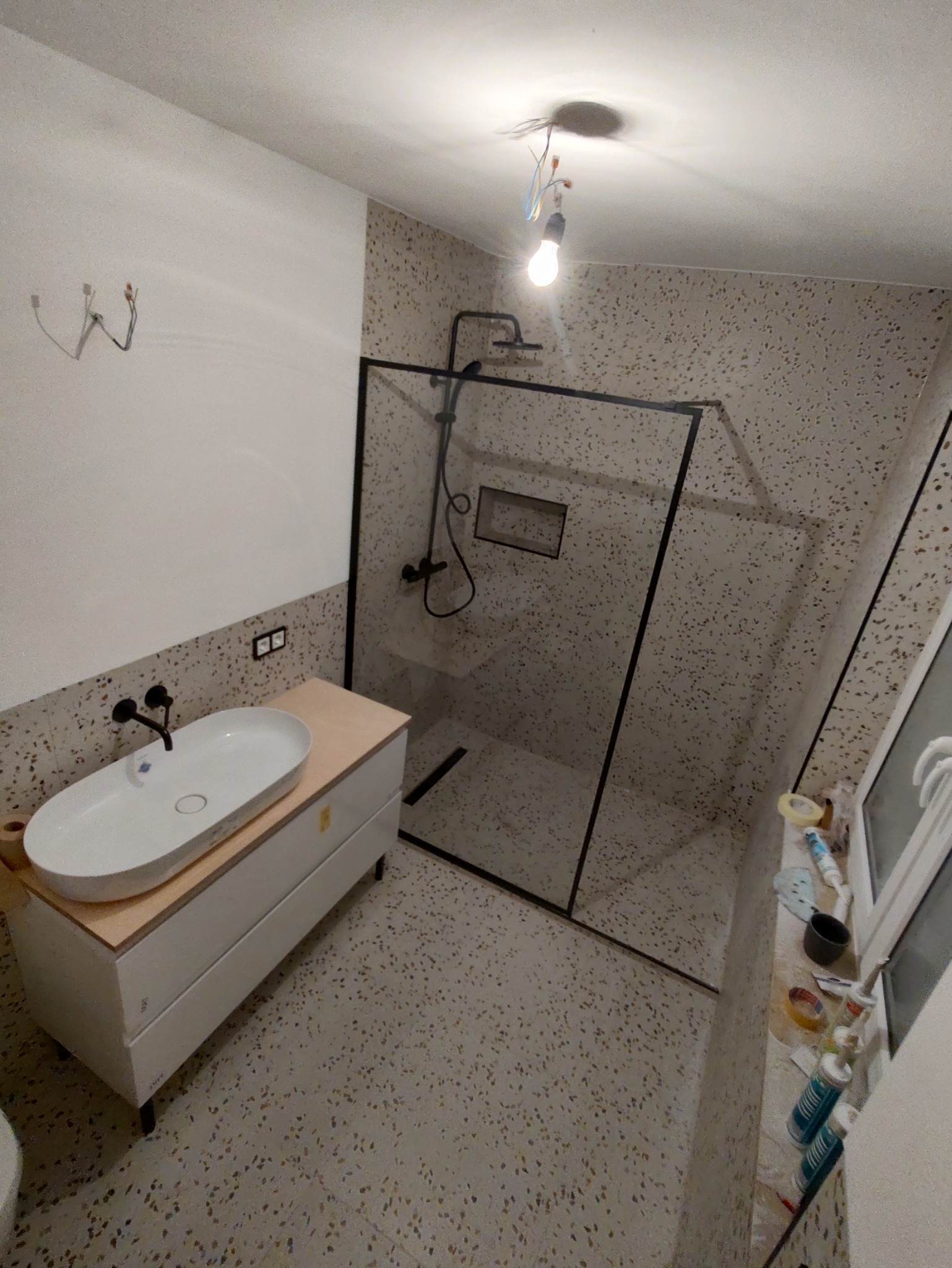 Nowoczesna łazienka z kabiną prysznicową typu walk-in, umywalką nablatową i płytkami lastryko na ścianach i podłodze. Widoczne niedokończone podłączenie oświetlenia.