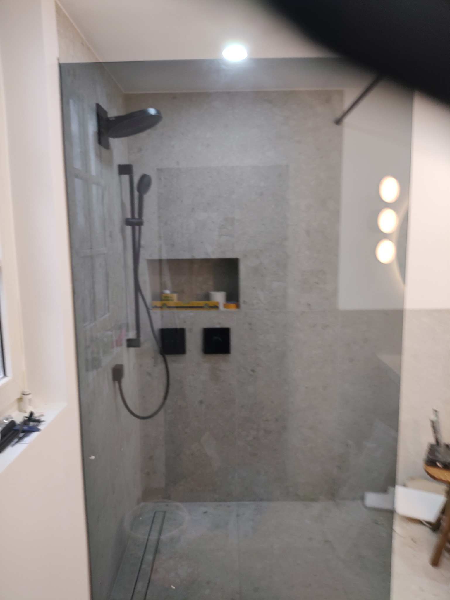 Nowoczesna kabina prysznicowa walk-in z szarymi płytkami imitującymi kamień, czarną armaturą i wnęką z półkami. Minimalistyczny design, oświetlenie punktowe.