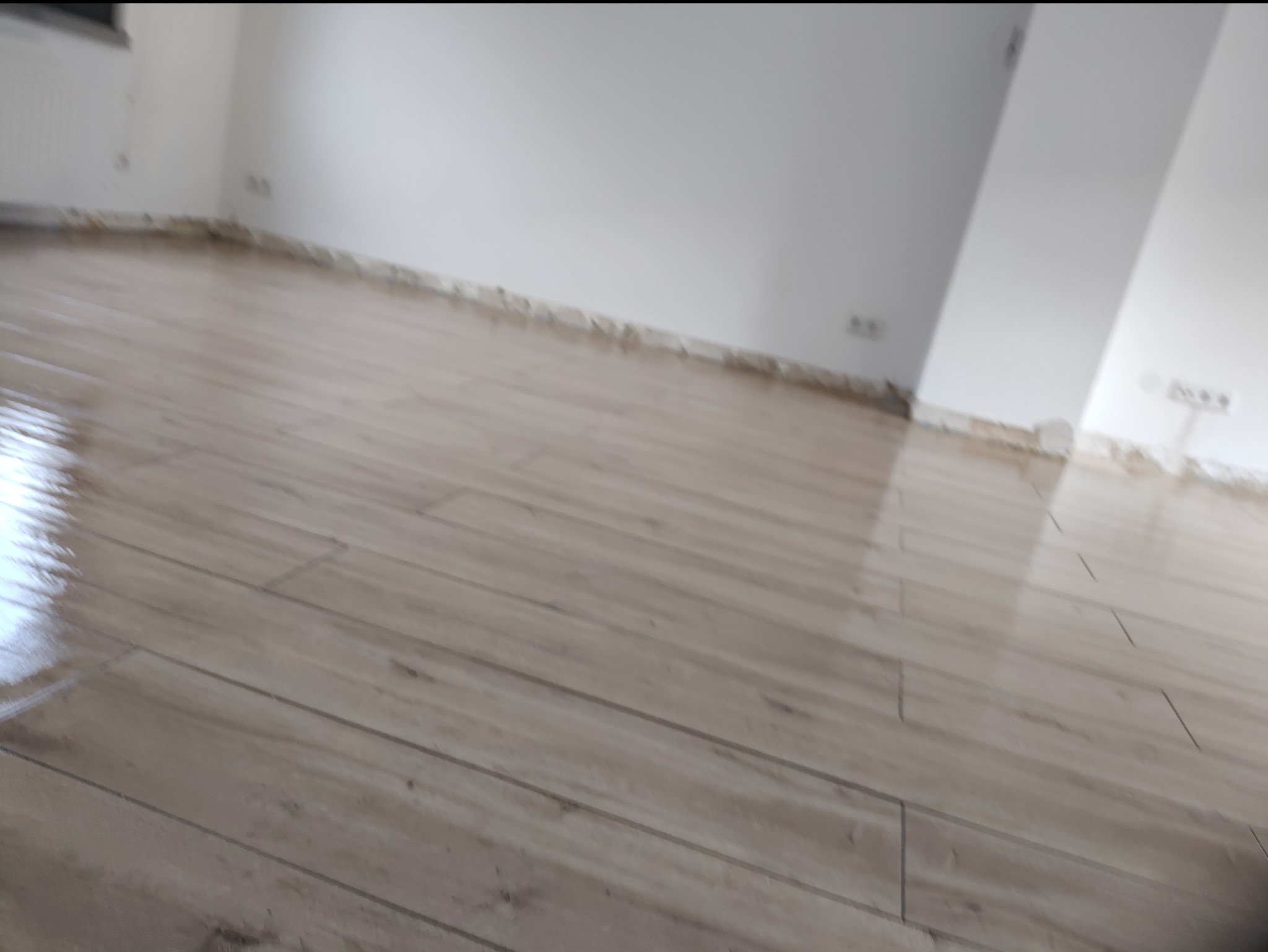 Ułożona podłoga z paneli imitujących drewno w jasnym wnętrzu. Widoczne ślady po remoncie przy ścianach. Nowa podłoga w trakcie wykańczania.