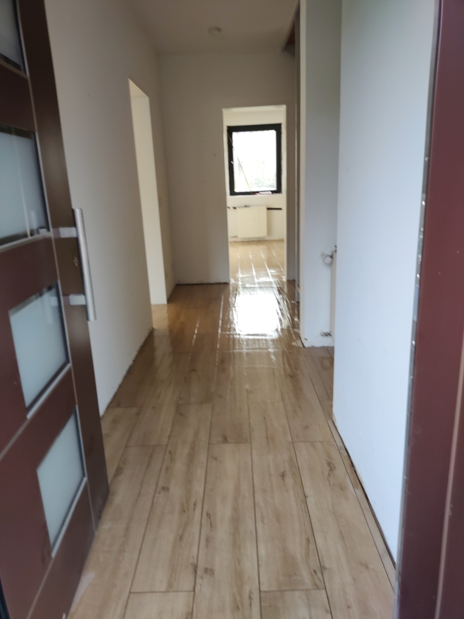 Korytarz z nową, jasną podłogą imitującą drewno, widok zza otwartych drzwi. Białe ściany, okno w tle. Podłoga błyszcząca, prawdopodobnie po umyciu.