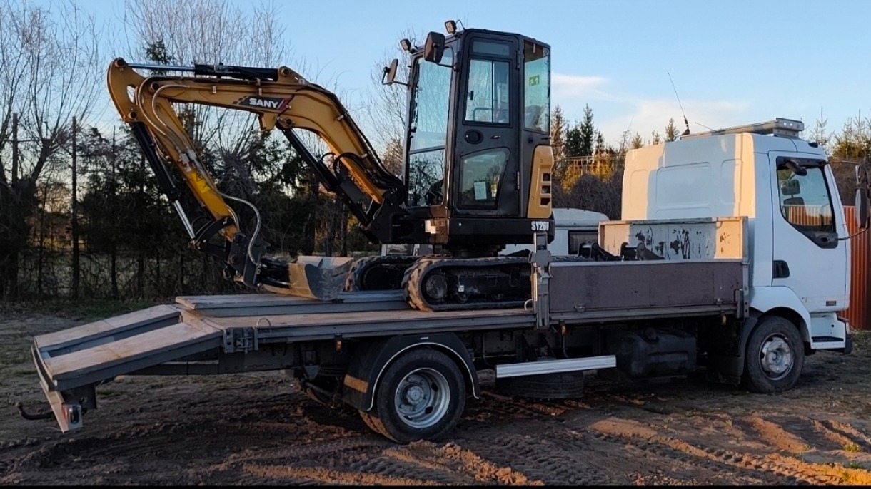 Żółty minikoparka SANY na platformie białej ciężarówki, gotowa do transportu. Widoczne opony i rampa załadunkowa. Tło: drzewa i błękitne niebo.