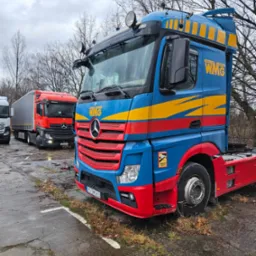 Niebiesko-czerwony ciągnik siodłowy WMG na placu, z widocznymi innymi ciężarówkami w tle i jesienną scenerią. Na kabinie żółte pasy i logo firmy.