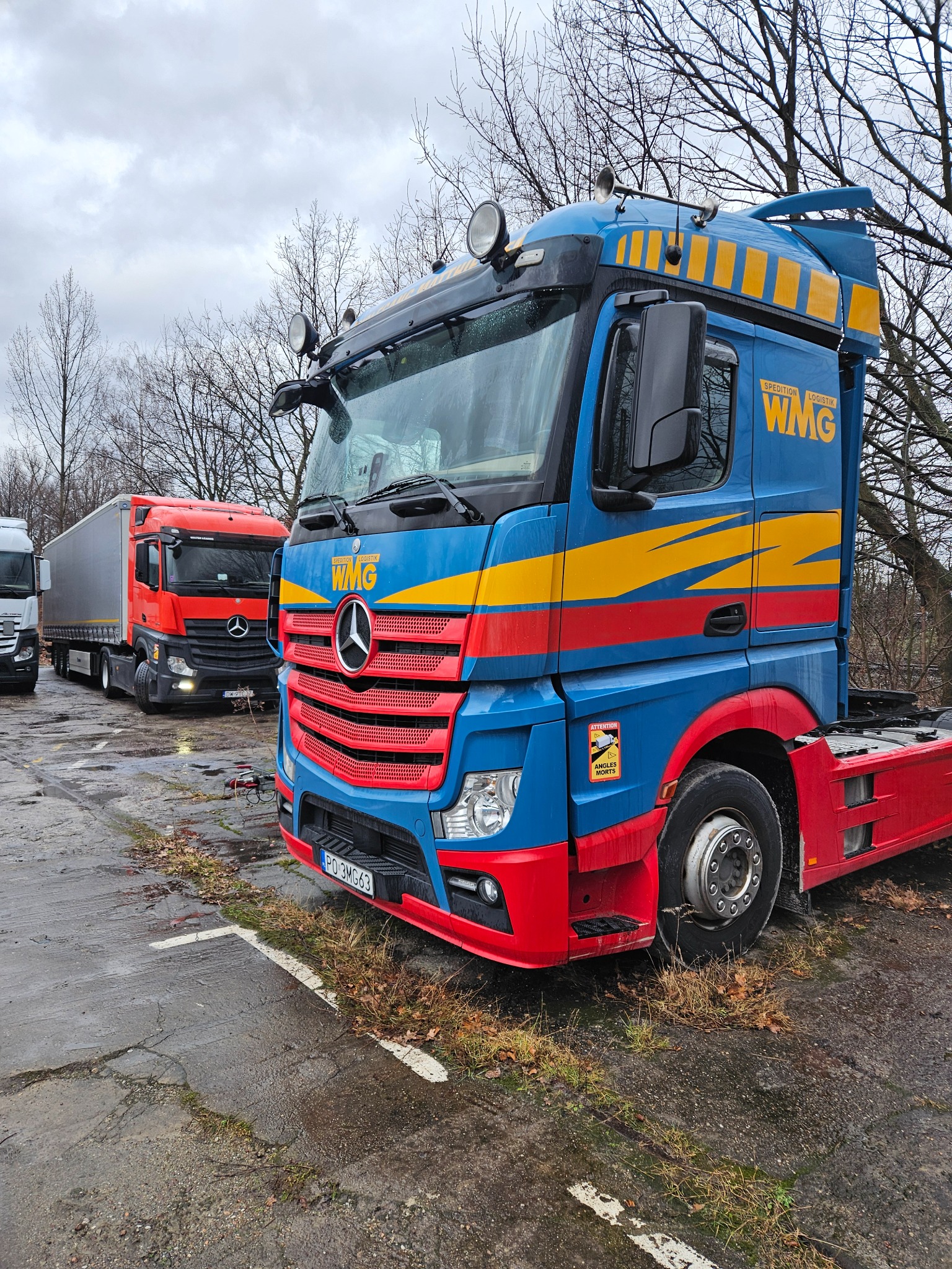 Niebiesko-czerwony ciągnik siodłowy WMG na placu, z widocznymi innymi ciężarówkami w tle i jesienną scenerią. Na kabinie żółte pasy i logo firmy.