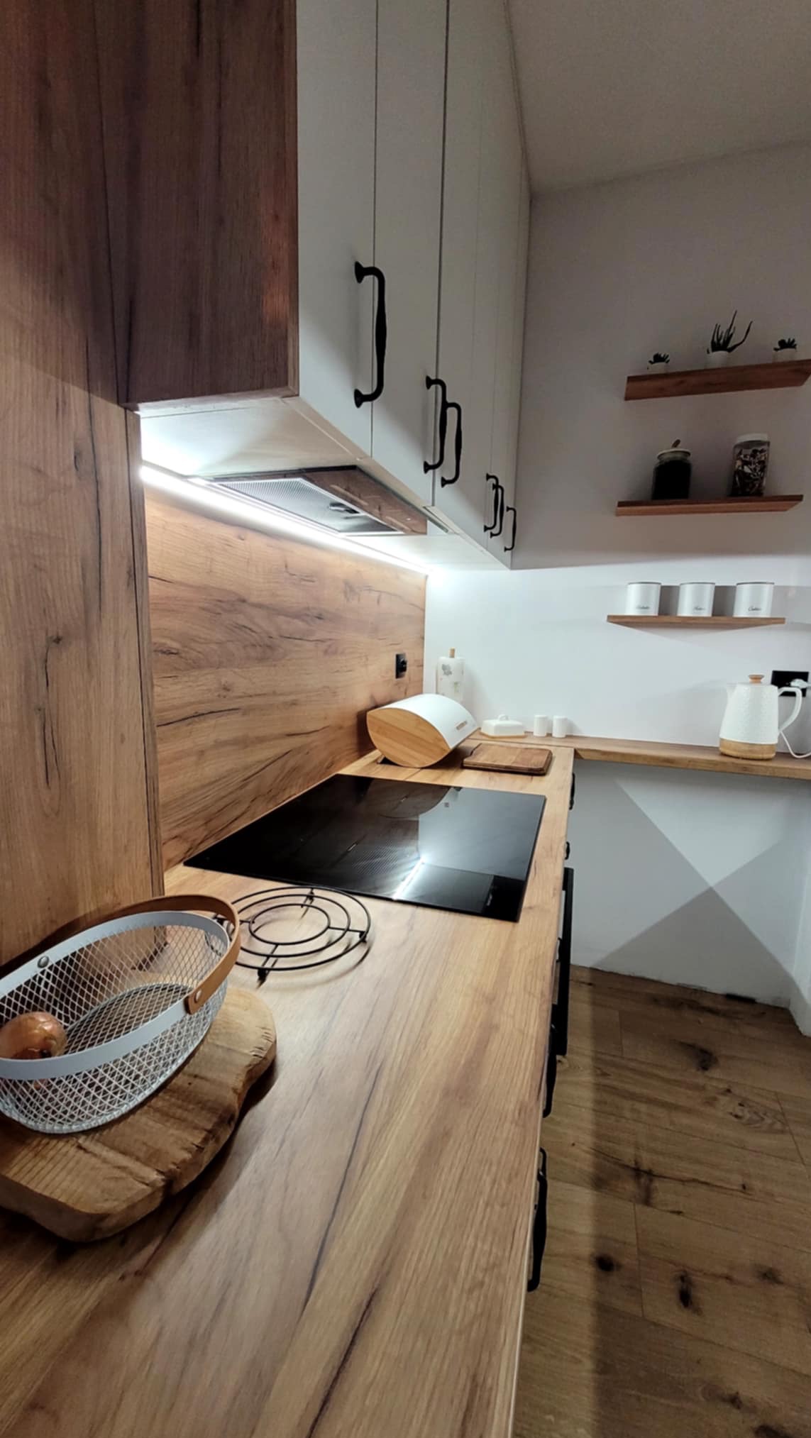 Nowoczesna kuchnia z drewnianymi frontami i białymi szafkami górnymi, czarne uchwyty. Blat z płyty imitującej drewno, oświetlenie LED pod szafkami.