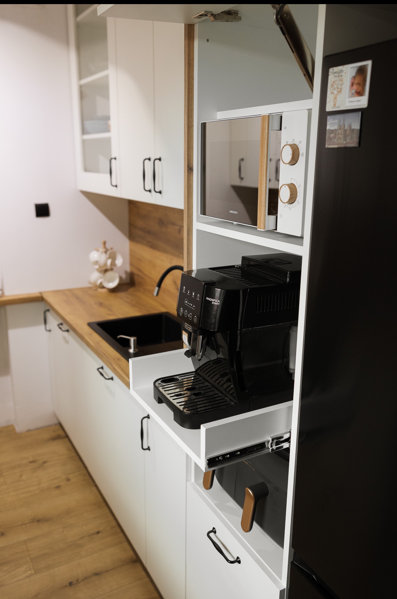 Biała zabudowa kuchenna z drewnianym blatem, czarnym zlewem i wbudowanym ekspresem do kawy na wysuwanej półce. Widoczna mikrofalówka i lodówka.
