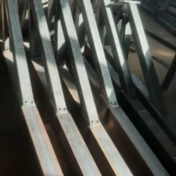 Metalowe konstrukcje kratownicowe, przygotowane do dalszego montażu, widoczne spawy i otwory montażowe, ułożone równolegle w warsztacie.