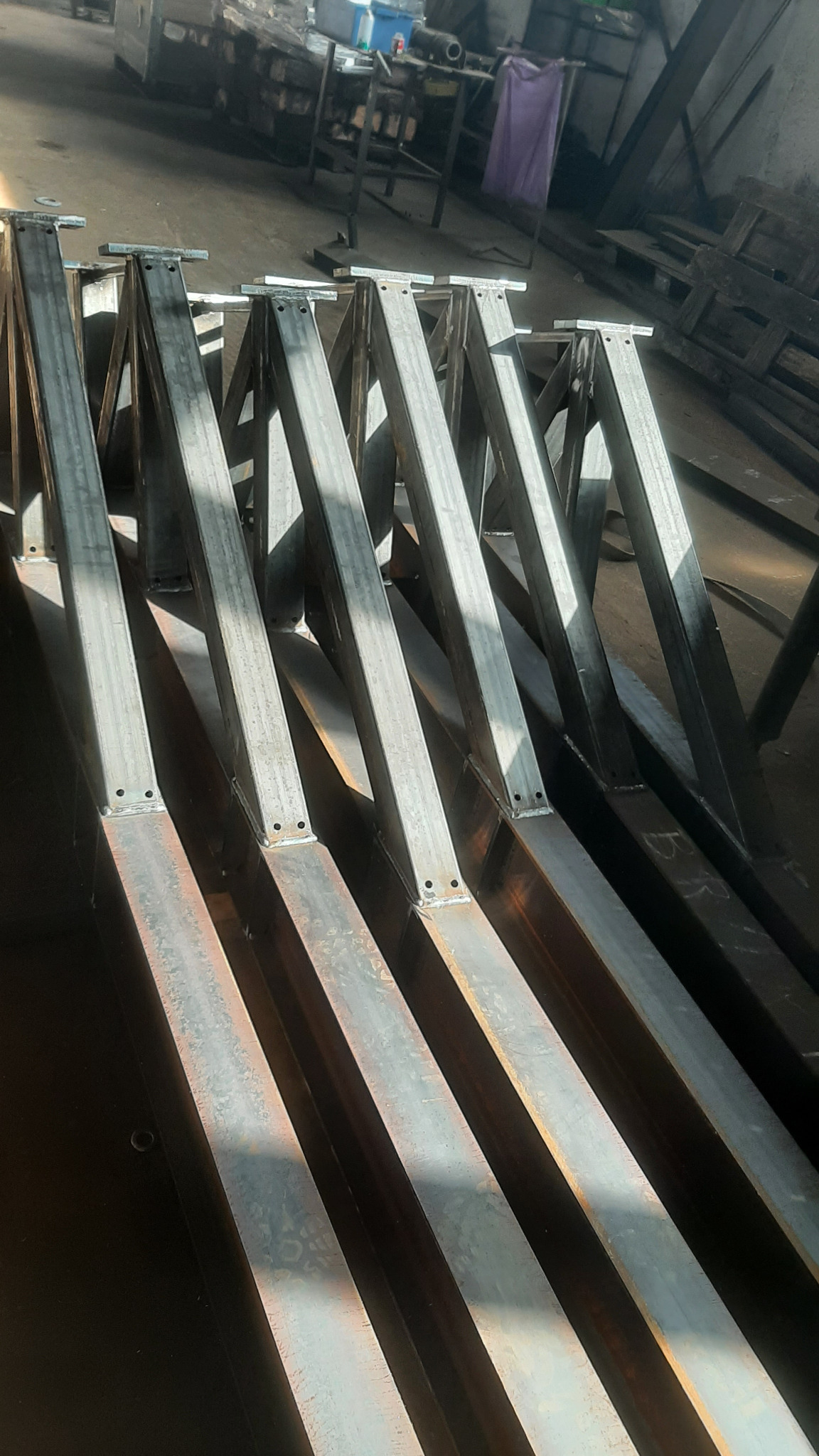 Metalowe konstrukcje kratownicowe, przygotowane do dalszego montażu, widoczne spawy i otwory montażowe, ułożone równolegle w warsztacie.