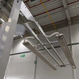 Metalowa konstrukcja z lampami w hali produkcyjnej. Widoczne przewody, elementy mocujące. Ujęcie z dołu, oświetlenie sufitowe.